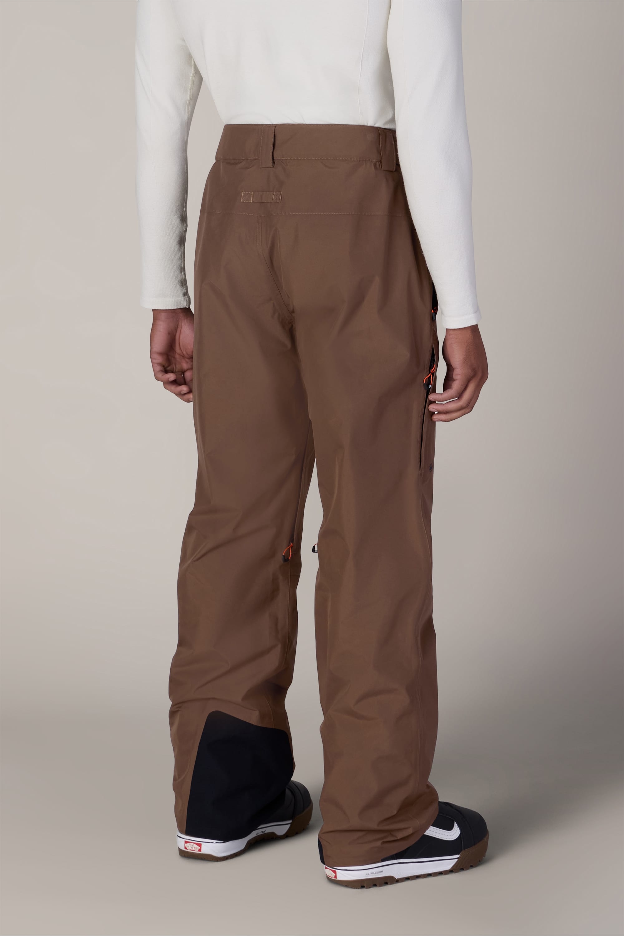 A person wearing the 686 Men’s GORE-TEX® Core Shell Pant by 686, paired with a white long-sleeve shirt and black shoes with white soles, stands with their back to the camera against a neutral background.