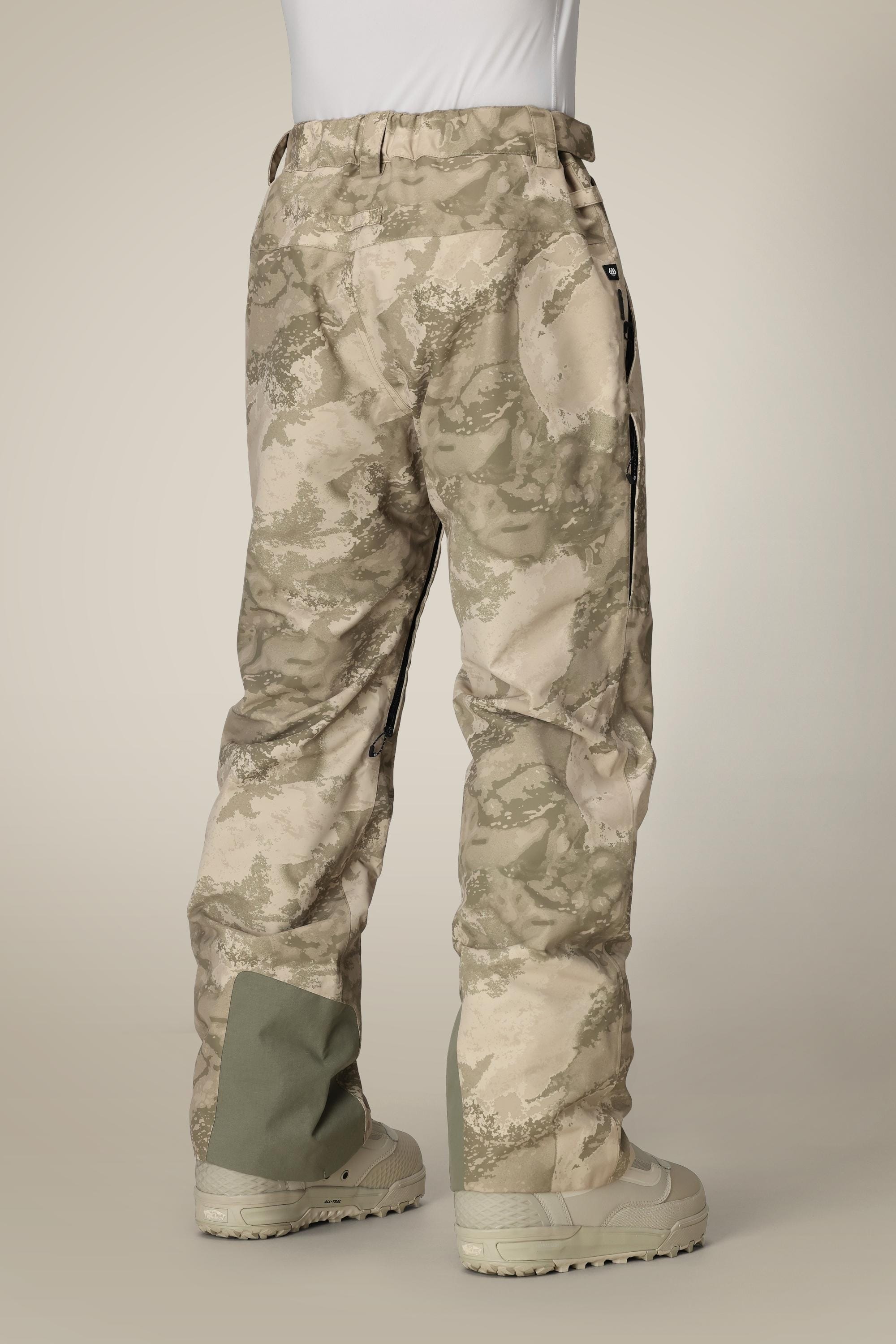 A person wearing 686 Mens GORE-TEX Core Shell Pant in light tan and brown, with pockets and reinforced green fabric on the lower legs, a white shirt tucked in, and beige boots, shown from the back.