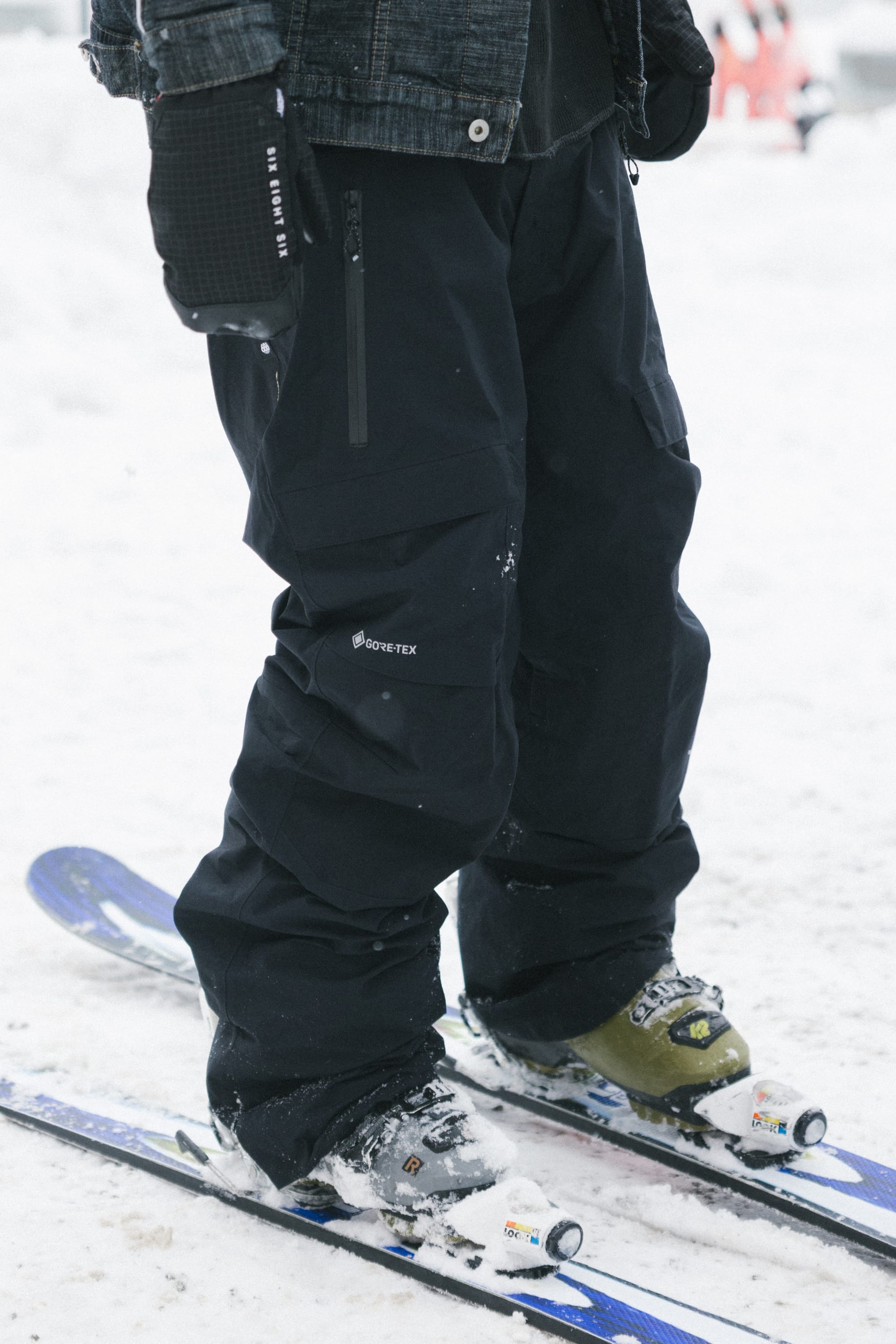 Person wearing 686 Men’s GORE-TEX® Dispatch™ Shell Pant by 686, a jacket, and gloves, standing on blue skis in the snow with green ski boots. The photo focuses on the lower half of their body.