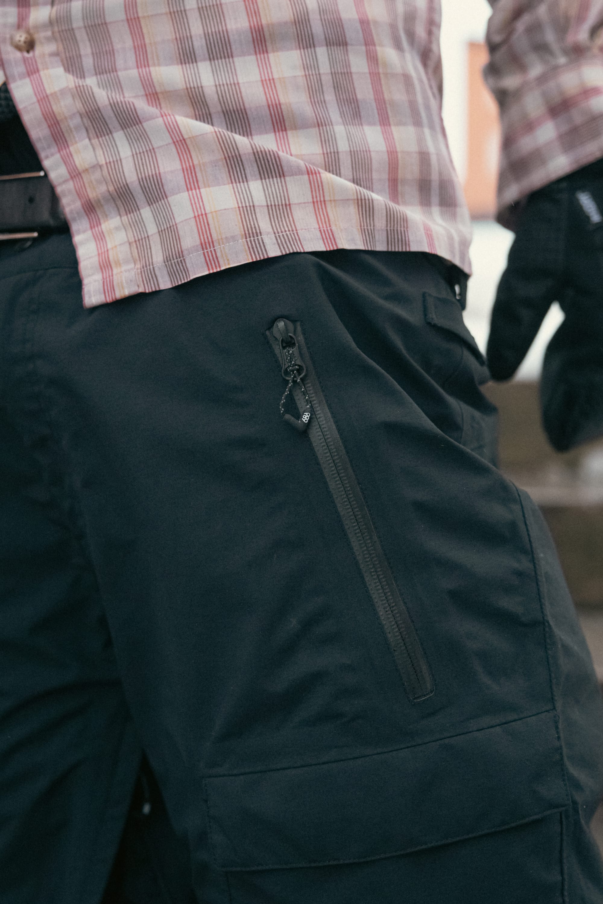 A person is shown from the waist down wearing 686 Men’s GORE-TEX® Dispatch™ Shell Pant, styled with black gloves and a plaid shirt; the pants feature a visible zippered pocket.