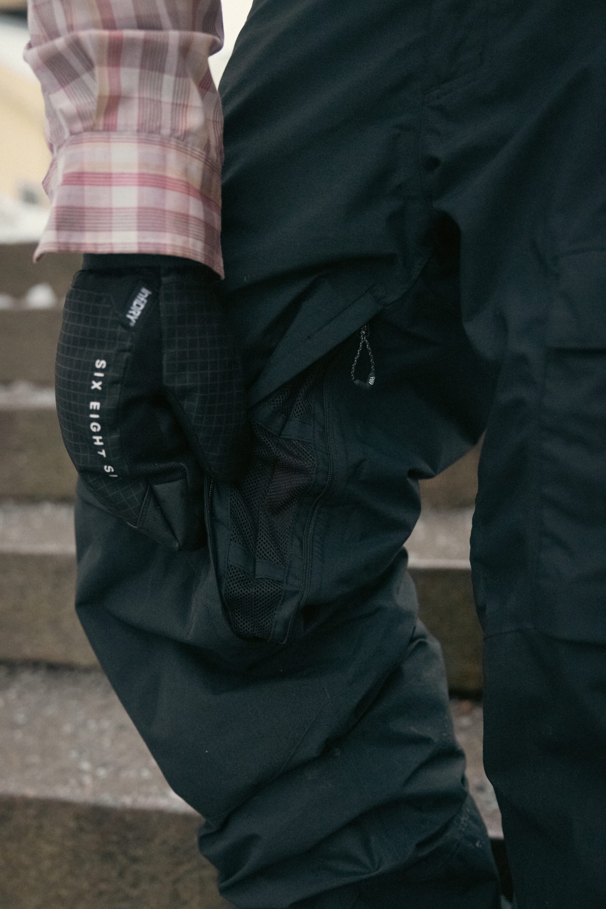 Wearing a plaid shirt, 686 Men’s GORE-TEX® Dispatch™ Shell Pant in black, and a black glove featuring SIX EIGHT SIX text, a person stands on steps with their gloved hand resting on their knee.