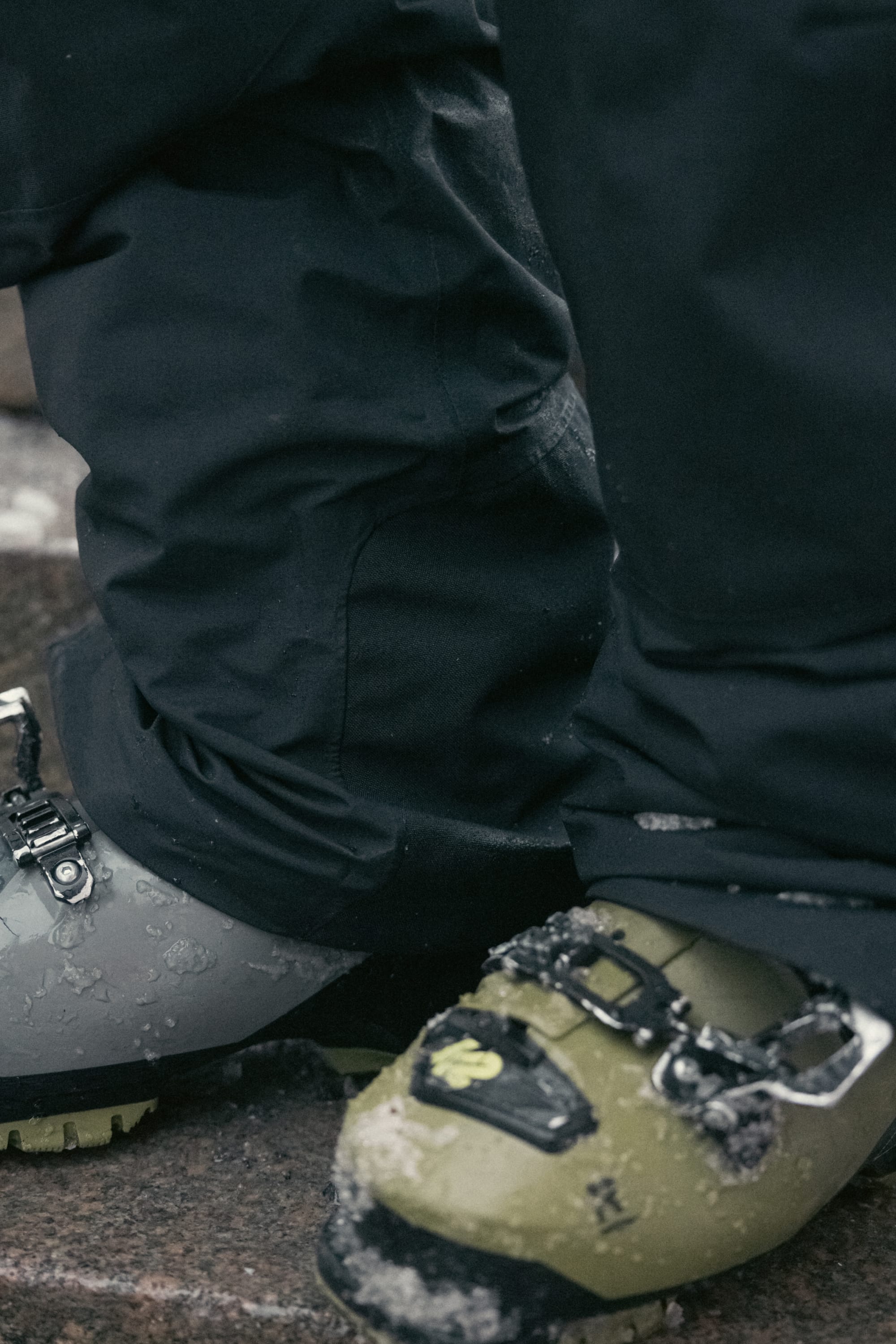 Close-up of a person wearing the 686 Men’s GORE-TEX® Dispatch™ Shell Pant and snow-covered green ski boots with buckles, standing on a snowy surface.