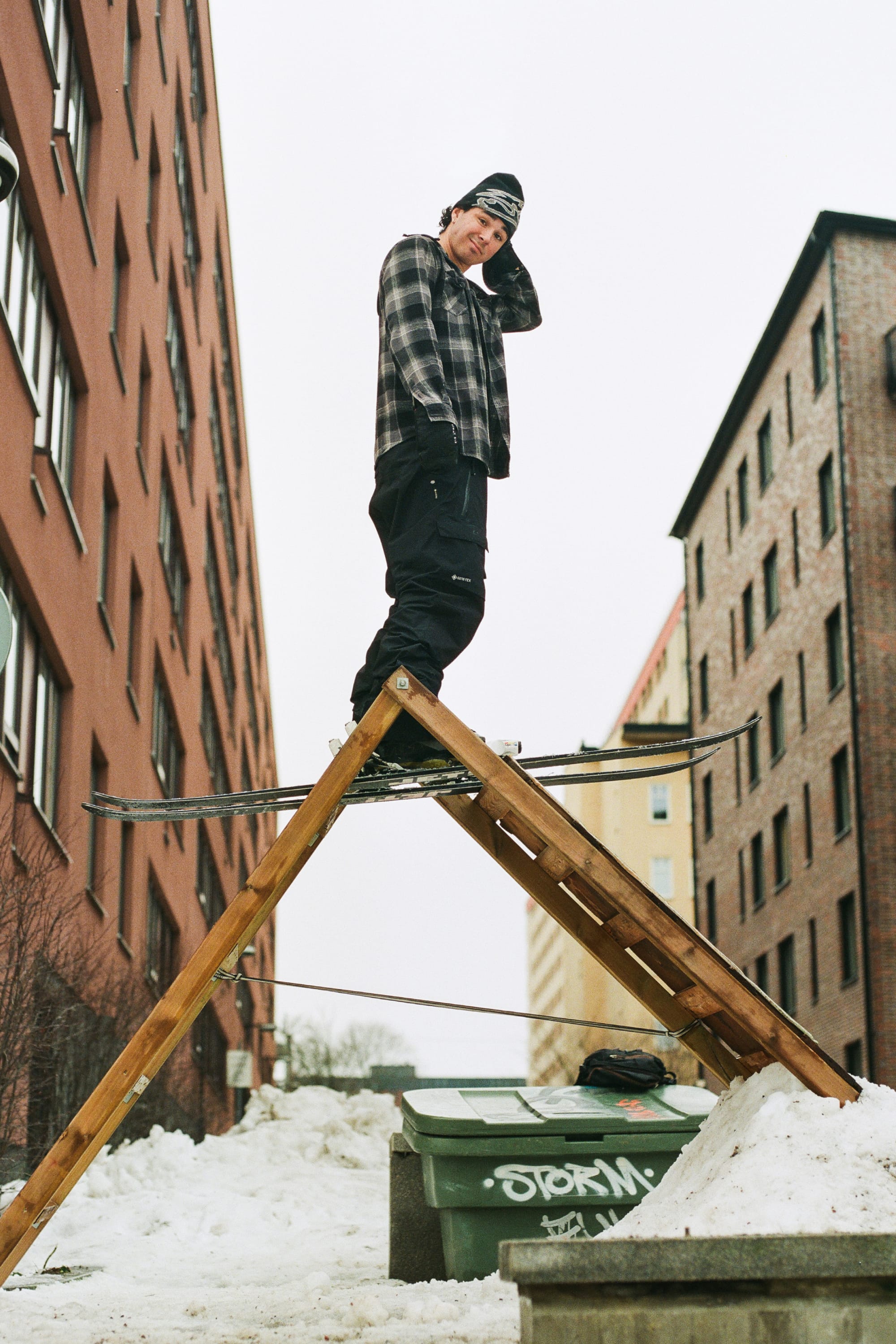Wearing the 686 Men’s GORE-TEX® Dispatch™ Shell Pant, a person stands on skis atop a wooden ramp in a snowy urban alleyway, framed by tall brick buildings.