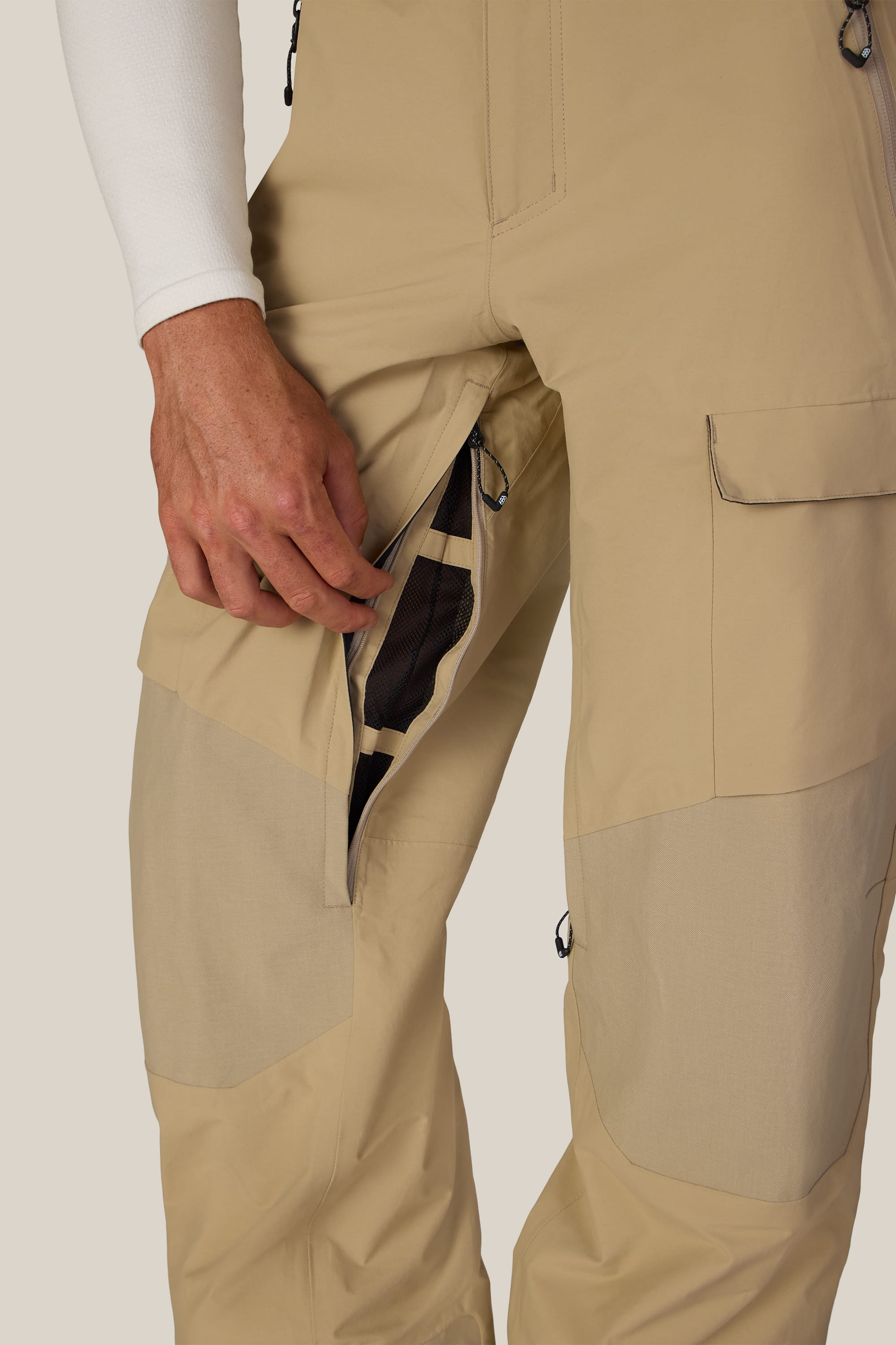 A person wearing 686 Men’s GORE-TEX® Dispatch™ Shell Pants in beige opens a zippered side pocket to reveal the mesh lining inside. Only the lower torso and hand are visible against a plain background.