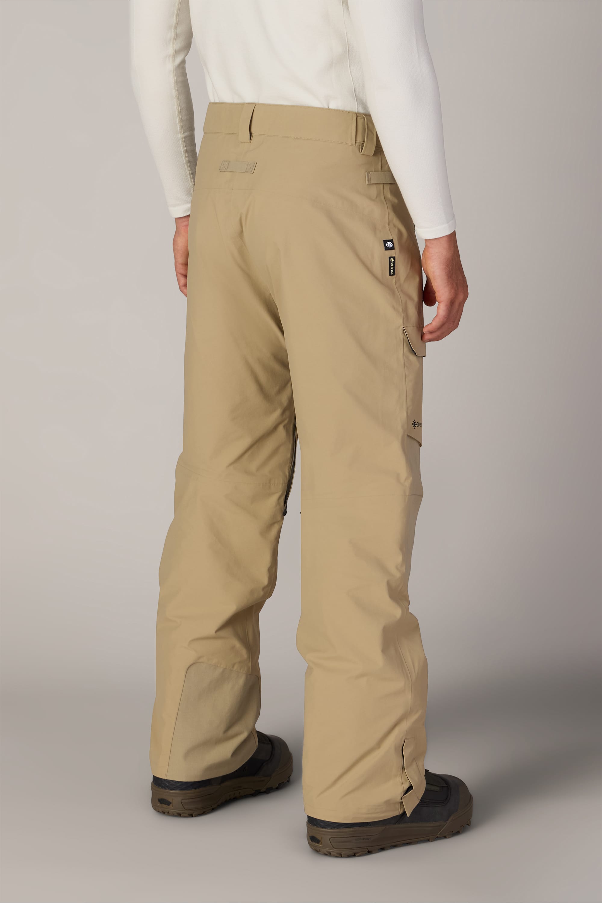 A person is seen from the back wearing the 686 Men’s GORE-TEX® Dispatch™ Shell Pant in tan, paired with black boots and a long-sleeve white shirt. The pants feature multiple pockets.