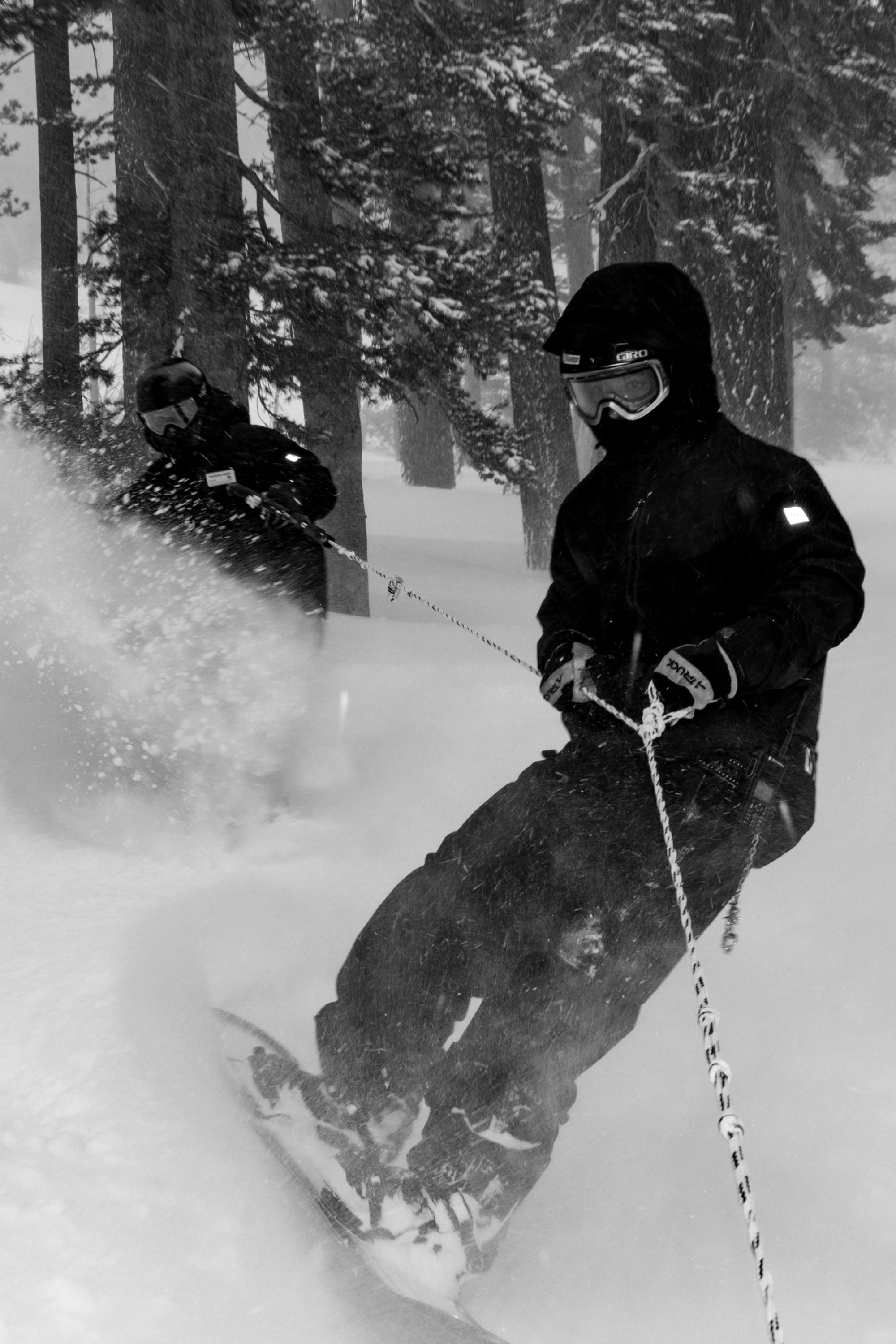 Wearing 686 Men’s GORE-TEX® Dispatch™ Shell Pants, two snowboarders in dark winter gear glide through deep forest snow, gripping ropes as snow sprays around them with tall trees rising in the background.
