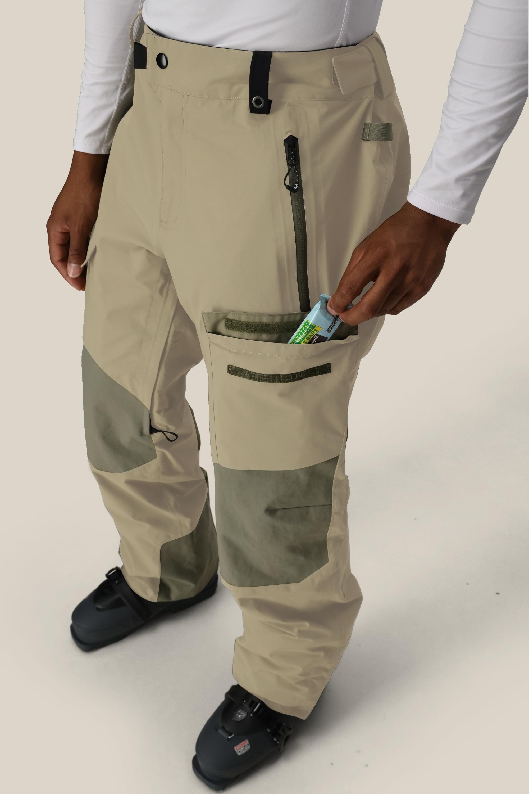 Wearing the 686 Men's GORE-TEX Dispatch™ Shell Pant in beige and olive green, along with black ski boots and a white long-sleeve shirt, a person puts a small packaged item into a zippered thigh pocket.