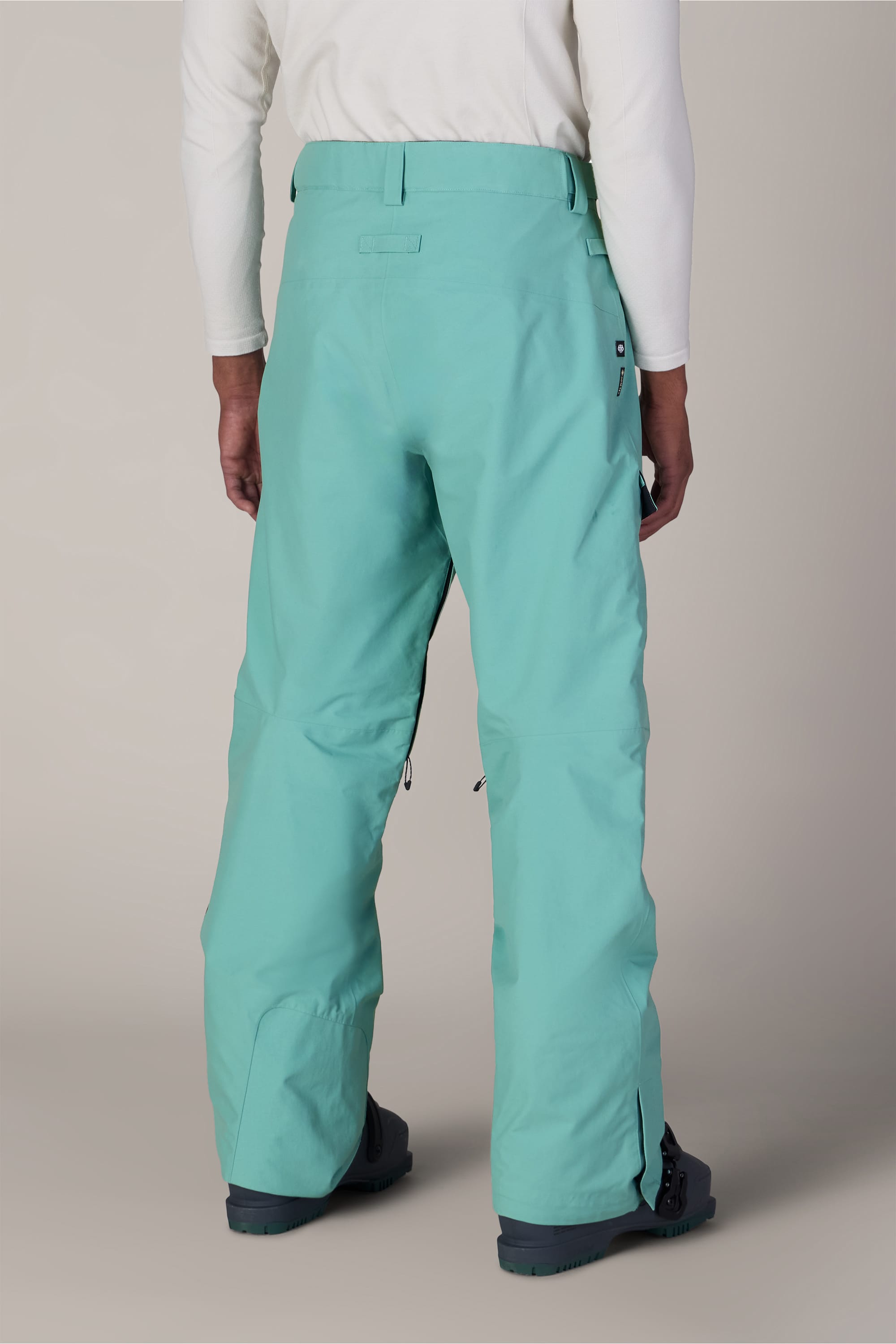 A person wearing 686 Men’s GORE-TEX® Dispatch™ Shell Pant in aqua blue, a white long-sleeve shirt, and black boots stands facing away against a neutral background.
