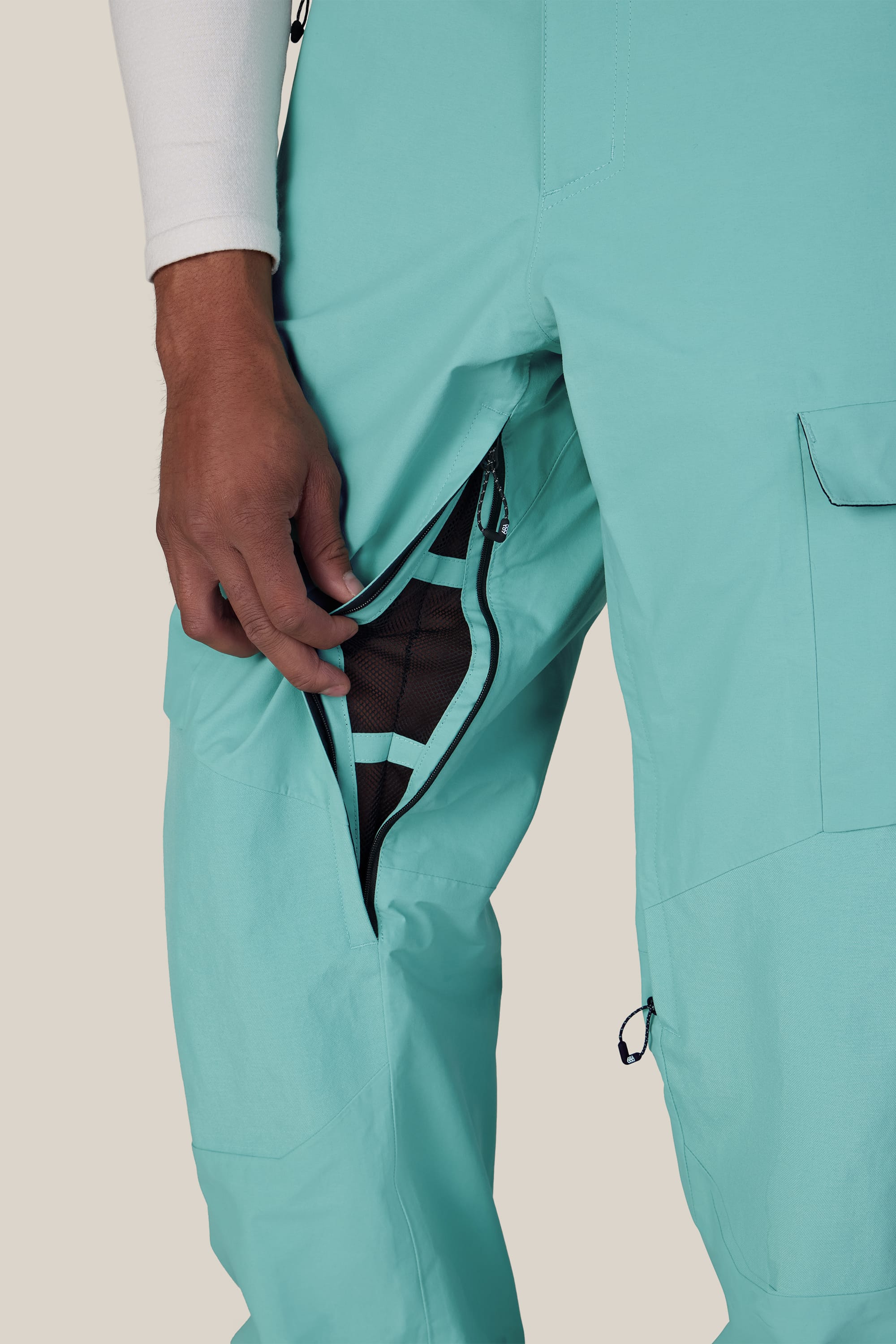 A person wearing the 686 Men’s GORE-TEX® Dispatch™ Shell Pant unzips a mesh-lined side vent, revealing breathable fabric underneath. Only their lower body and left hand are visible against a plain background.