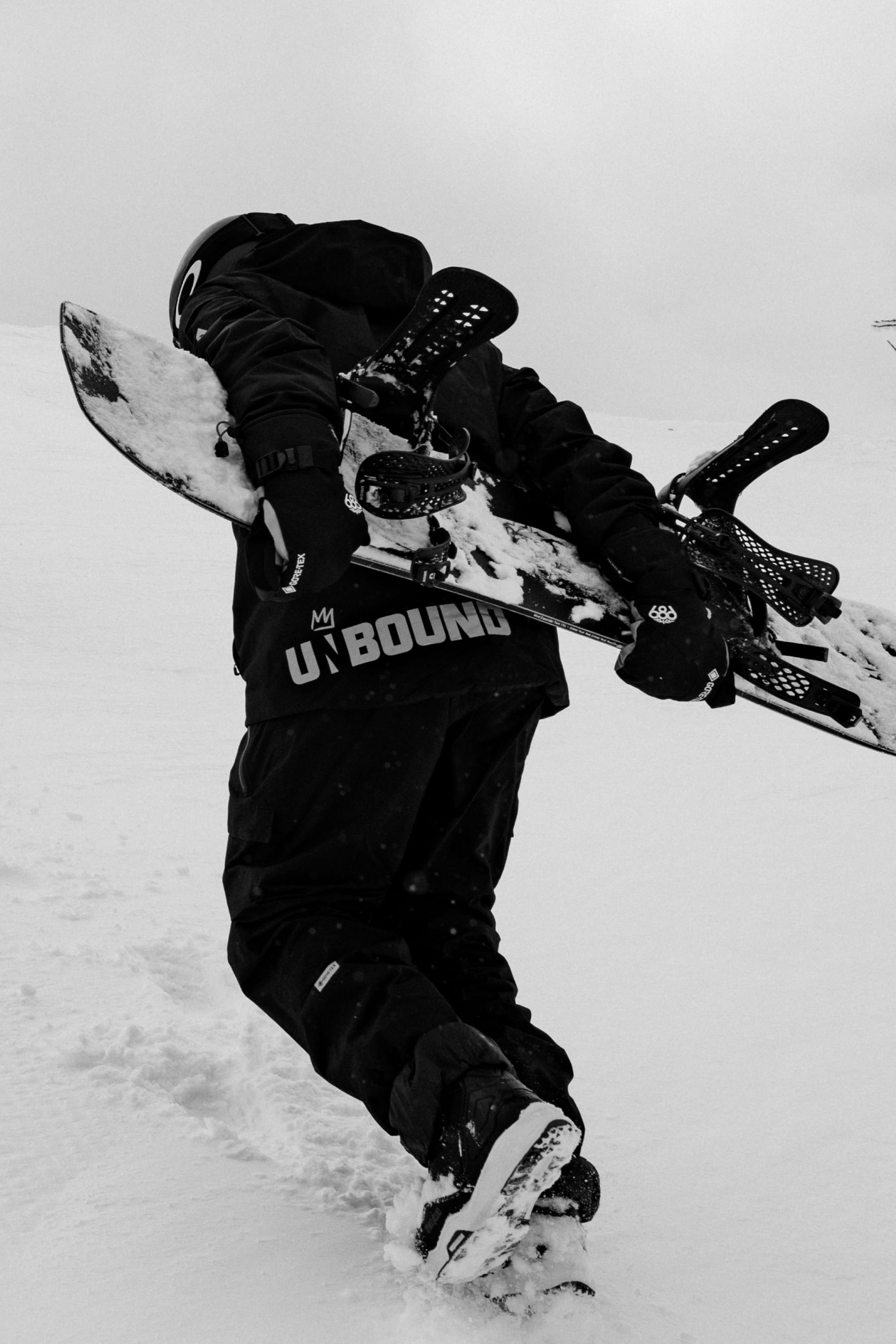 Clad in 686 Men’s GORE-TEX® Dispatch™ Shell Pants, a person in winter gear carries a snowboard up a snowy slope, seen from behind in black and white.