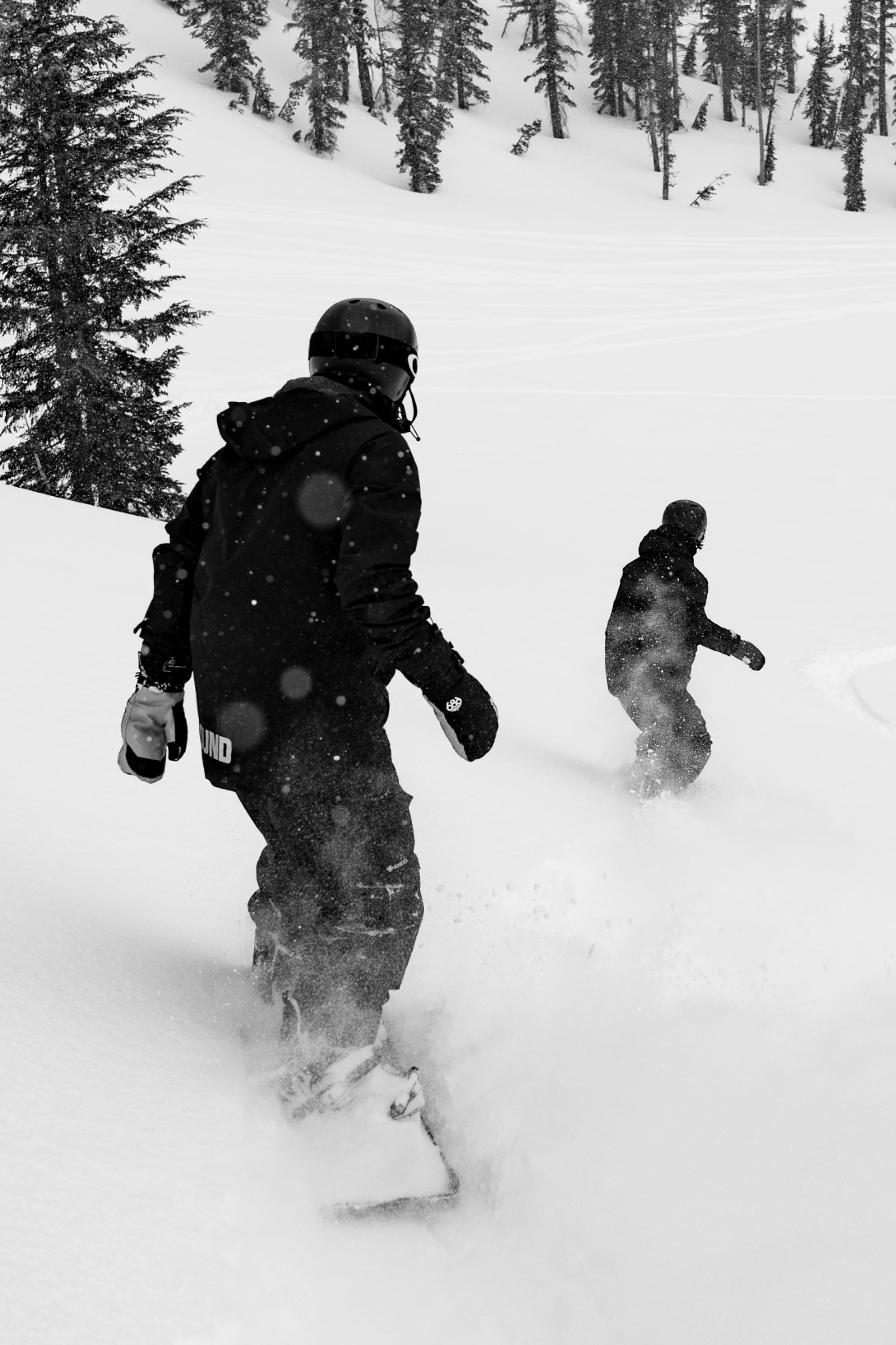 Wearing 686 Men’s GORE-TEX® Dispatch™ Shell Pants, two people snowboard down a snowy slope lined with trees. Snow sprays behind them as they ride, both outfitted in helmets and winter gear. The scene appears in black and white.