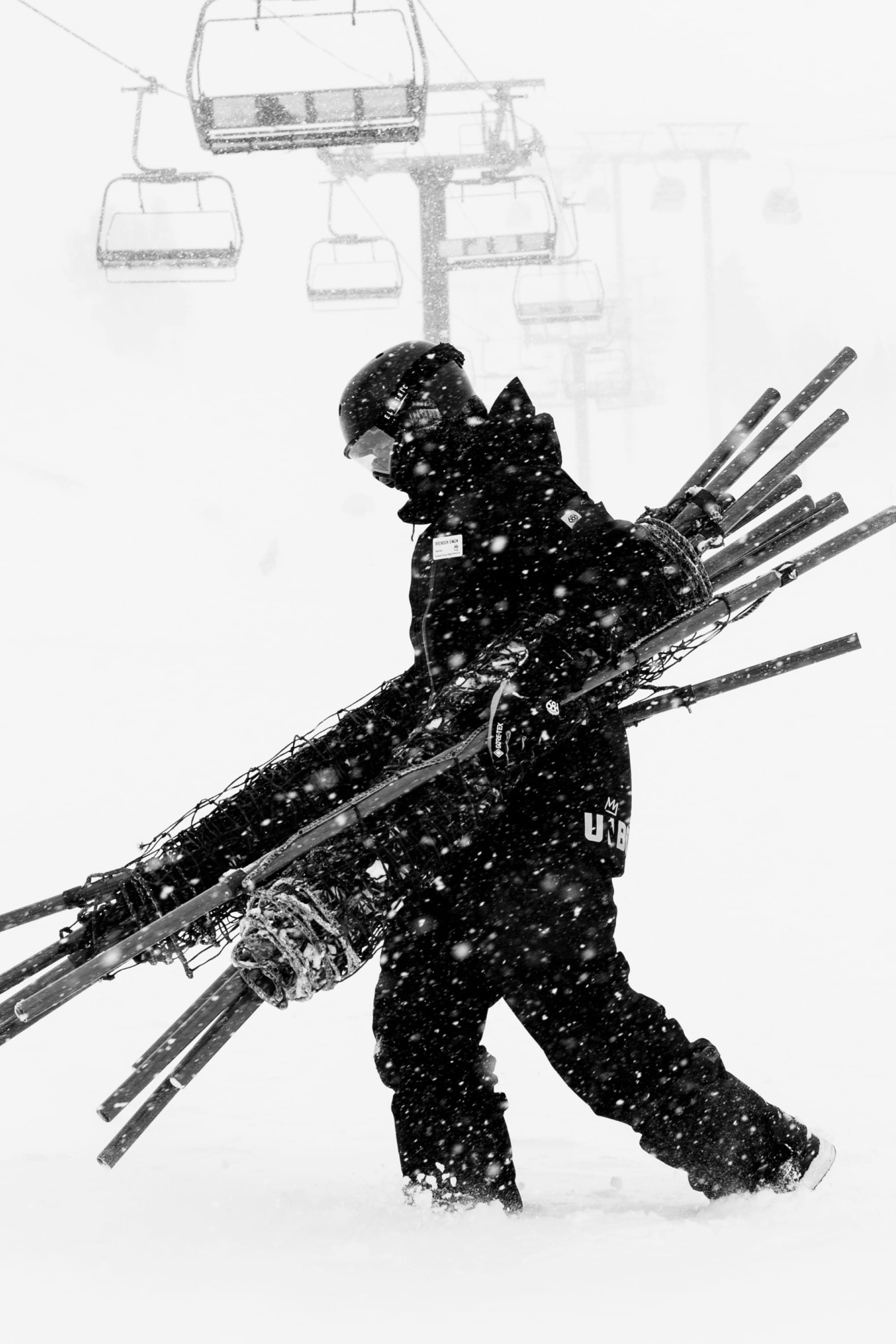 Wearing the 686 Men’s GORE-TEX® Dispatch™ Shell Pant, a person in winter gear carries ski poles and fencing through falling snow near empty ski lifts on a snowy mountain.