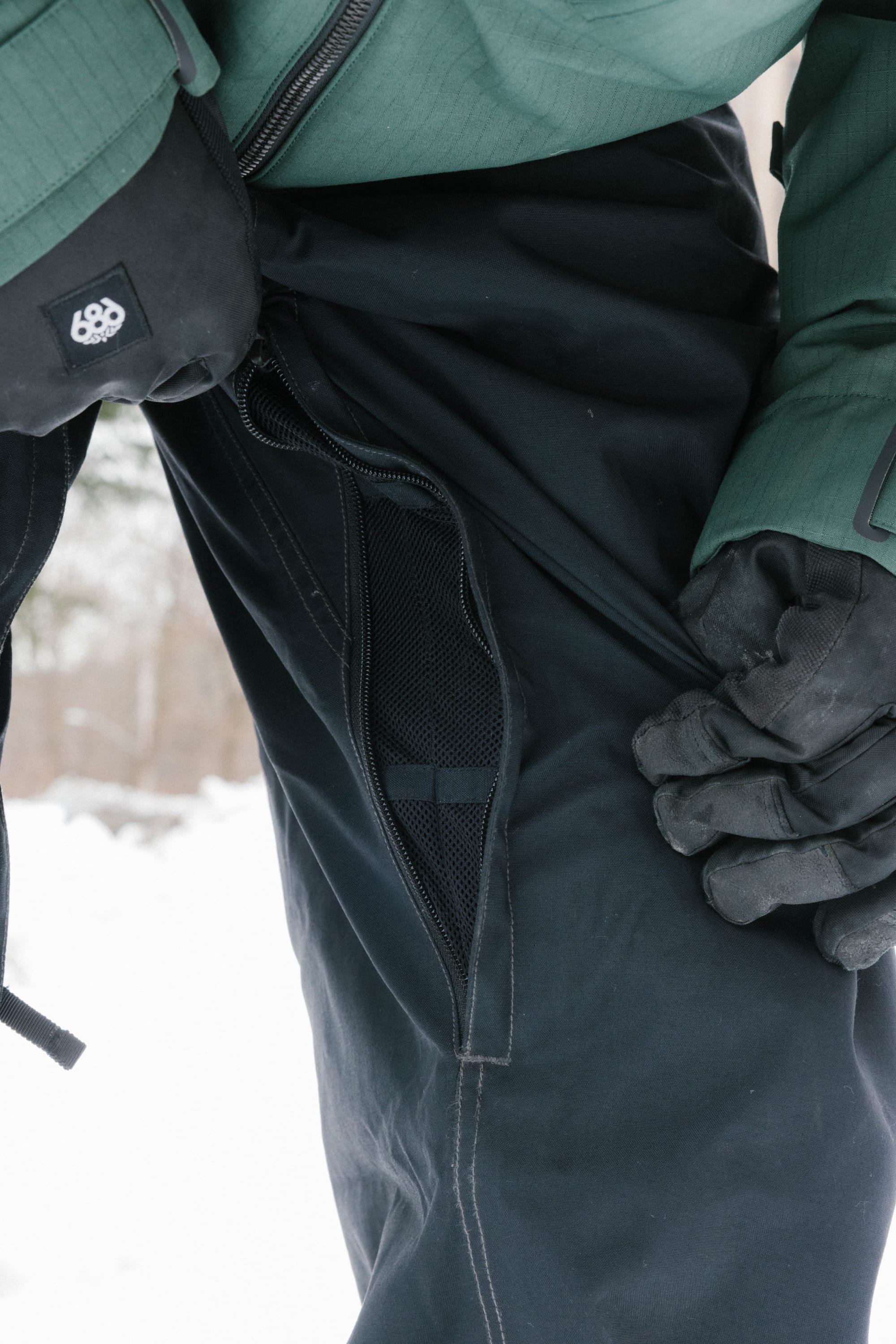 Wearing the 686 Dojo® Pant, a person in black nylon waterproof pants and green outerwear unzips a mesh-lined side vent. They're also wearing black gloves, with snow in the background.