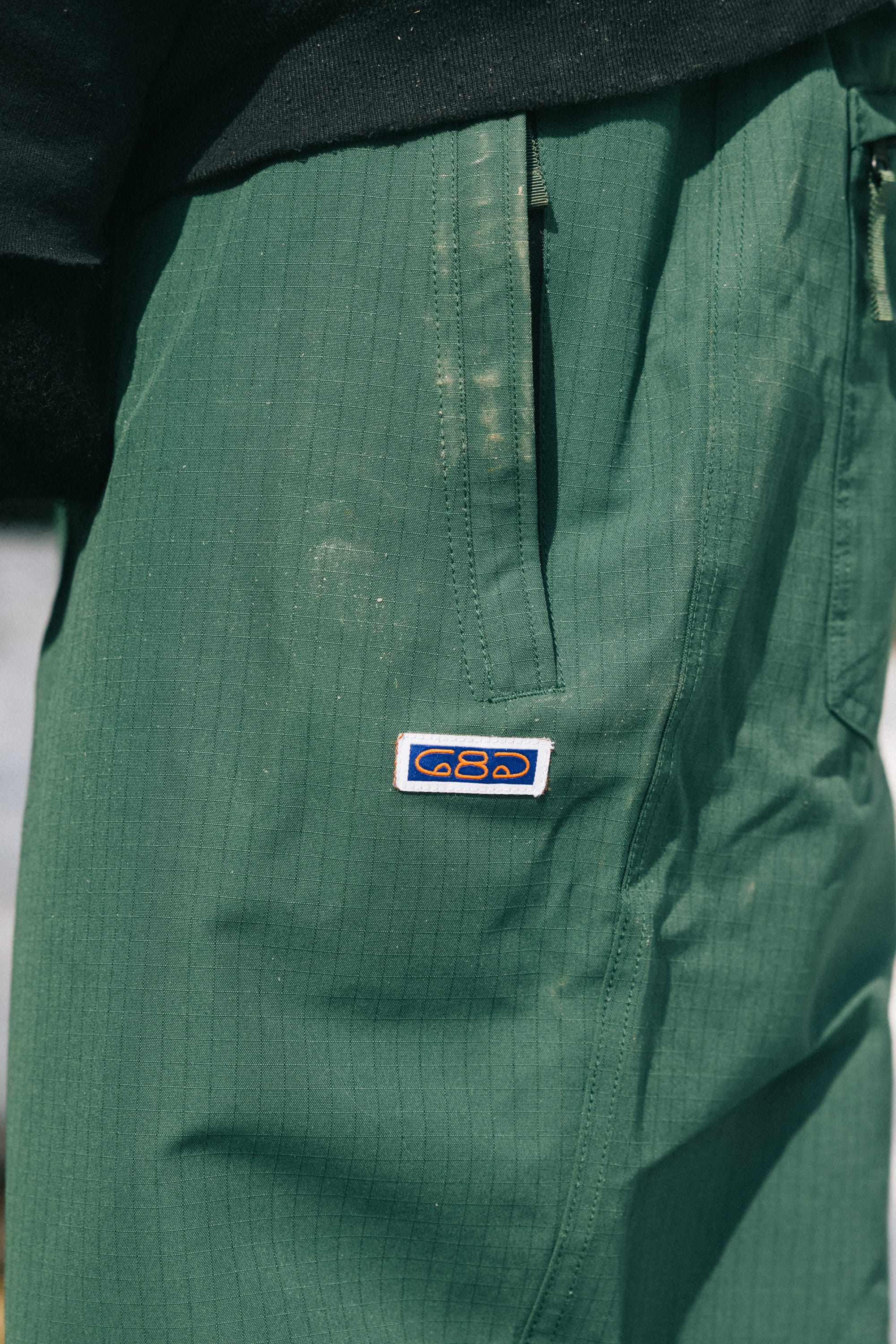 Close-up of a person wearing the 686 Dojo® Pant by 686 in green with a subtle grid pattern and an embroidered blue and white patch near the pocket. The person's black shirt is partially visible.