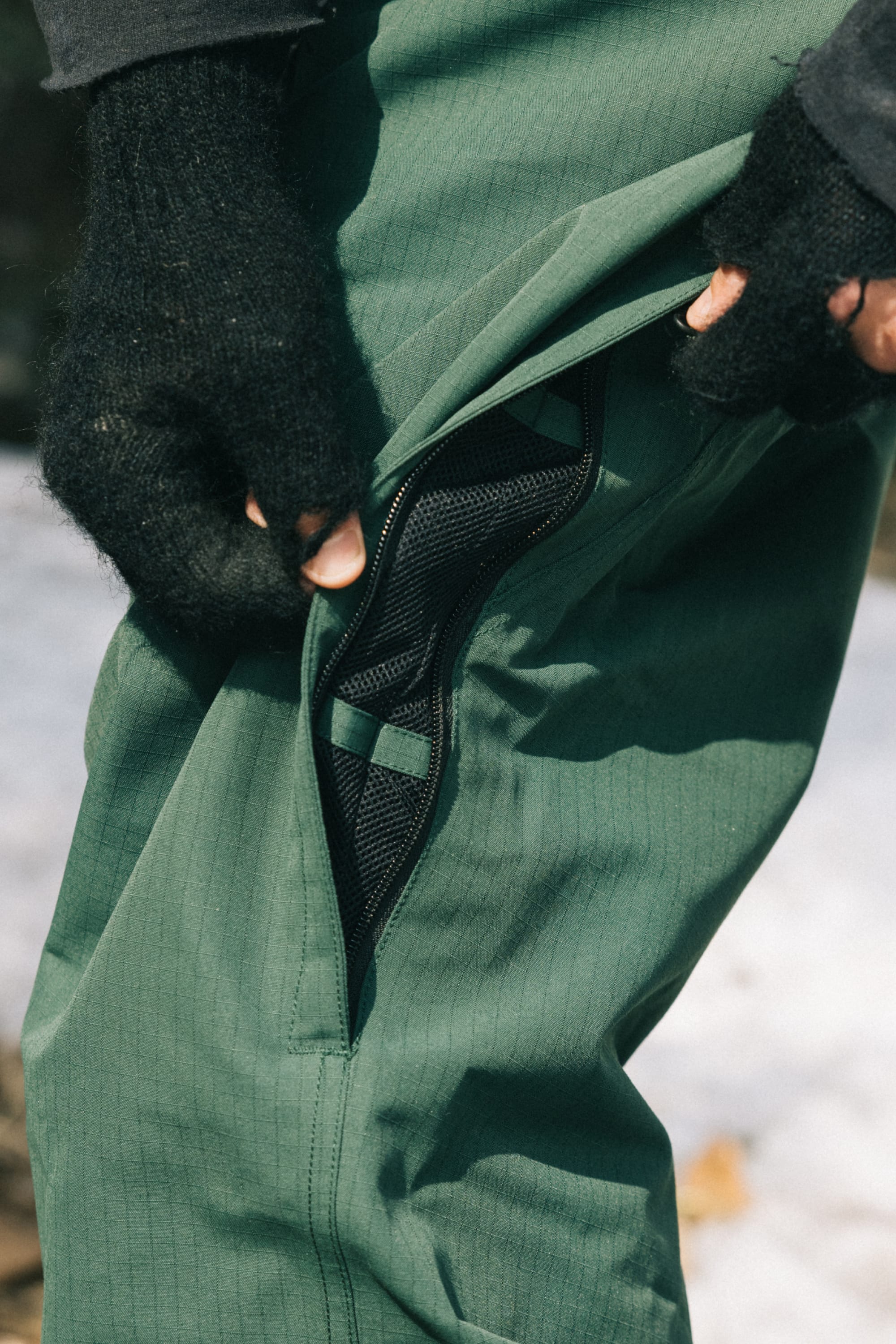 A person wearing 686 Dojo® Pant men's waterproof pants in green and black fingerless gloves opens a side zipper to reveal a mesh lining, with snow visible on the ground in the background.