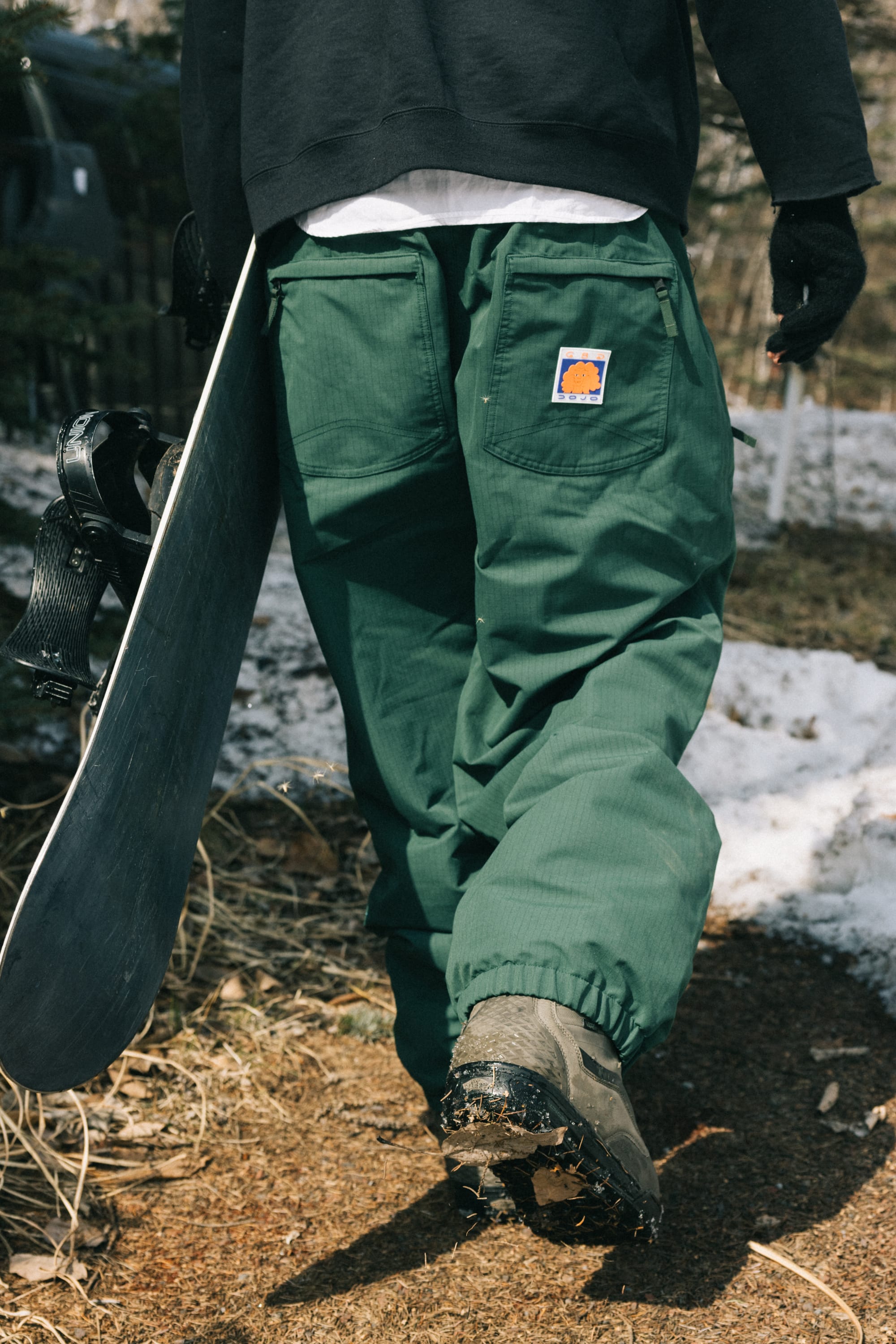Wearing 686 Dojo® Pants and boots, a person carries a snowboard down a muddy, snowy path. A sun logo patch is visible on the back pocket, and they stay warm with gloves and a black jacket.