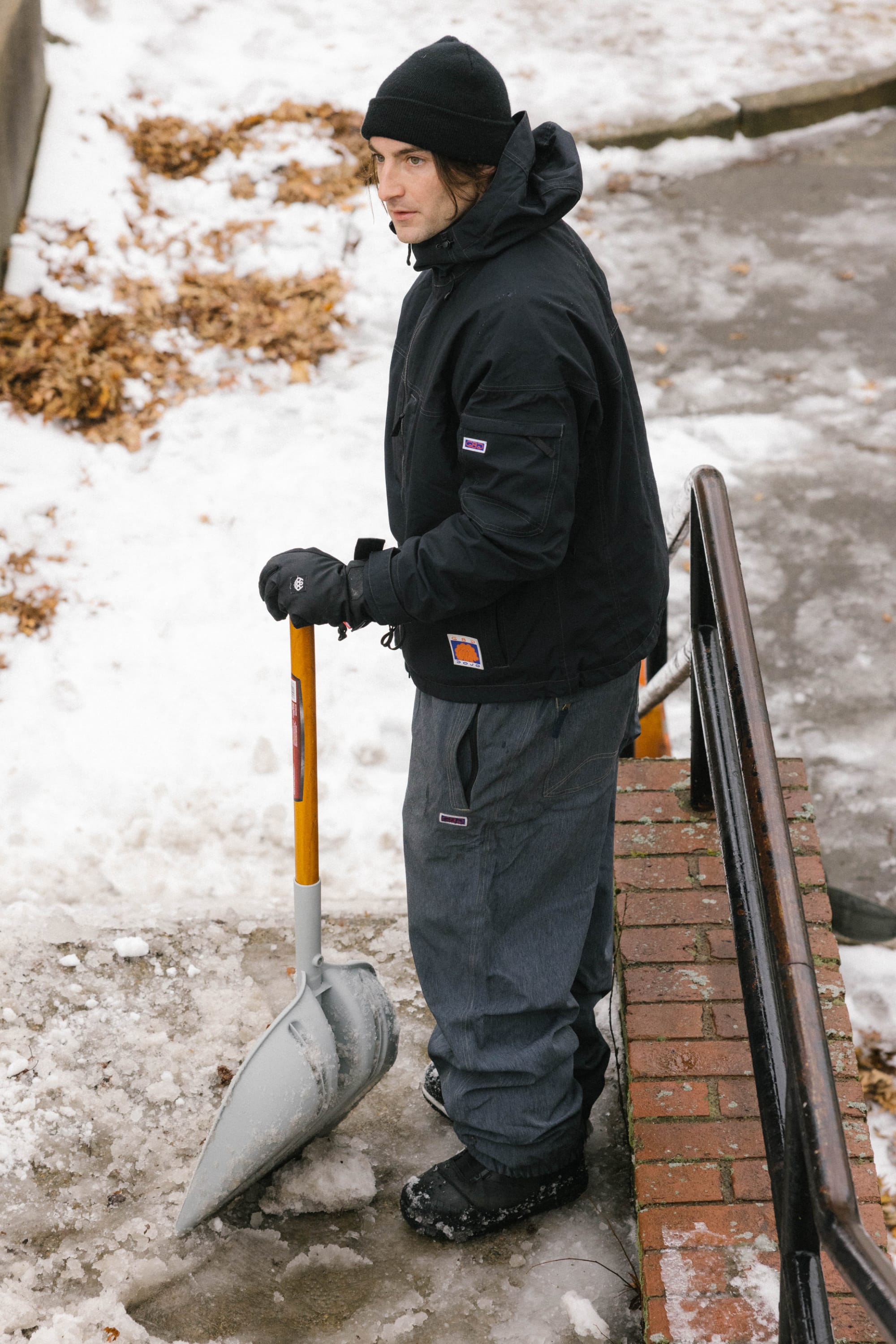 A person dressed in winter gear, including 686 Dojo® Pant by 686 and gloves, stands outdoors on an icy path holding a snow shovel near a brick railing, with patches of snow and ice scattered on the ground.