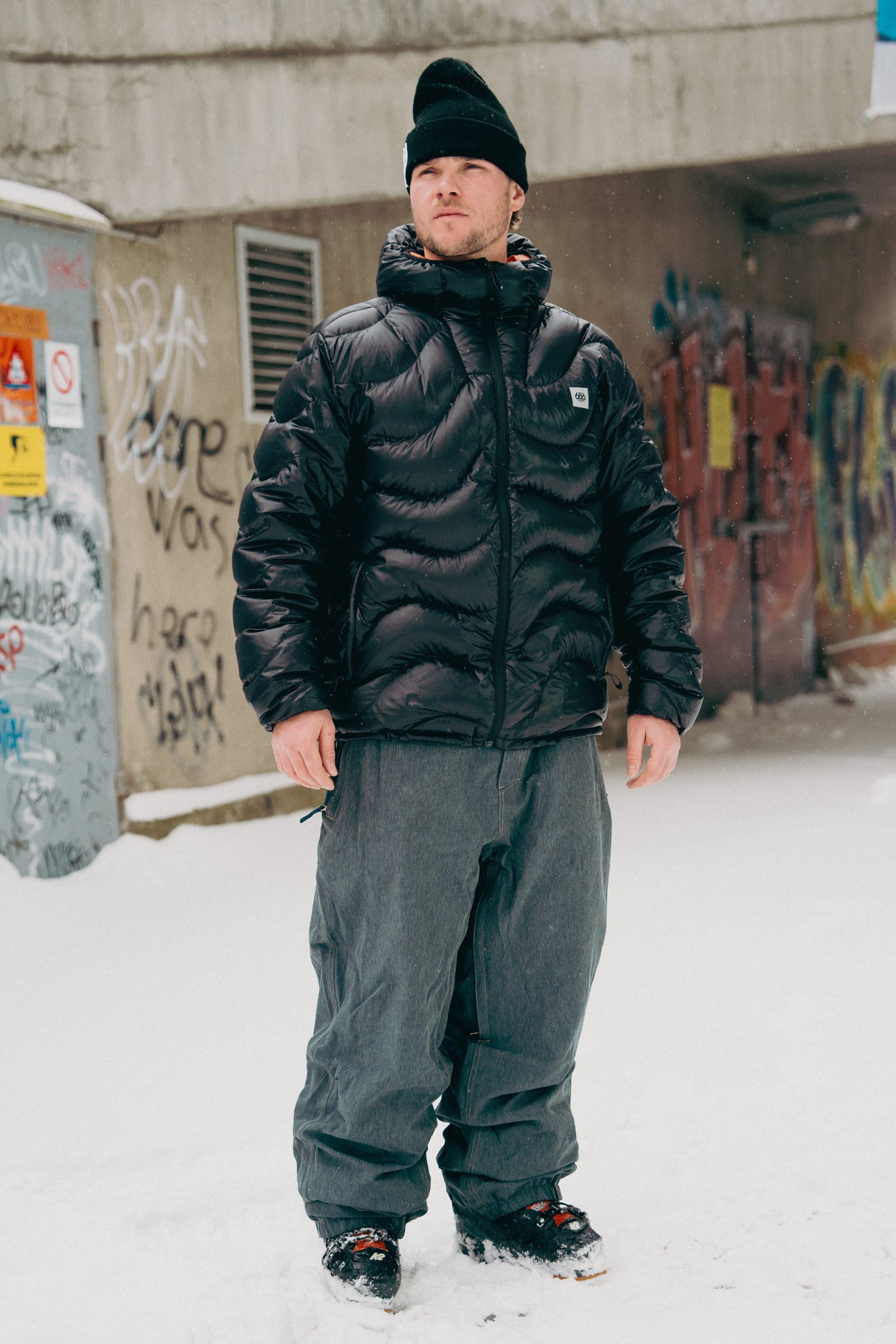 A man wearing a black puffer jacket, 686 Dojo® Pant by 686, black beanie, and black sneakers stands on snowy ground in front of graffiti-covered urban walls.