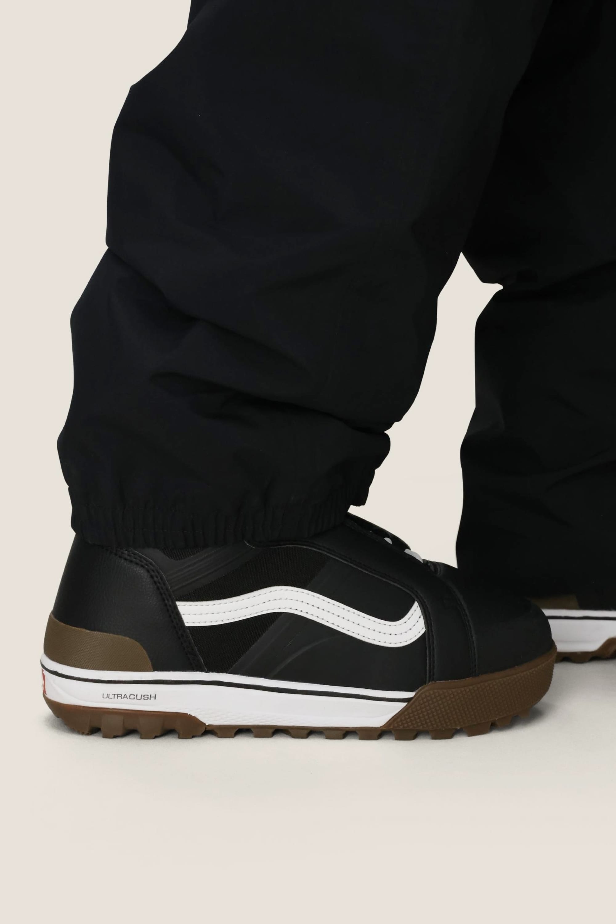Close-up of a person wearing black sneakers with white stripes, a brown sole, and the 686 Dojo® GORE-TEX Pant by 686, featuring men's woven fabric and elastic cuffs for a sporty, casual look.