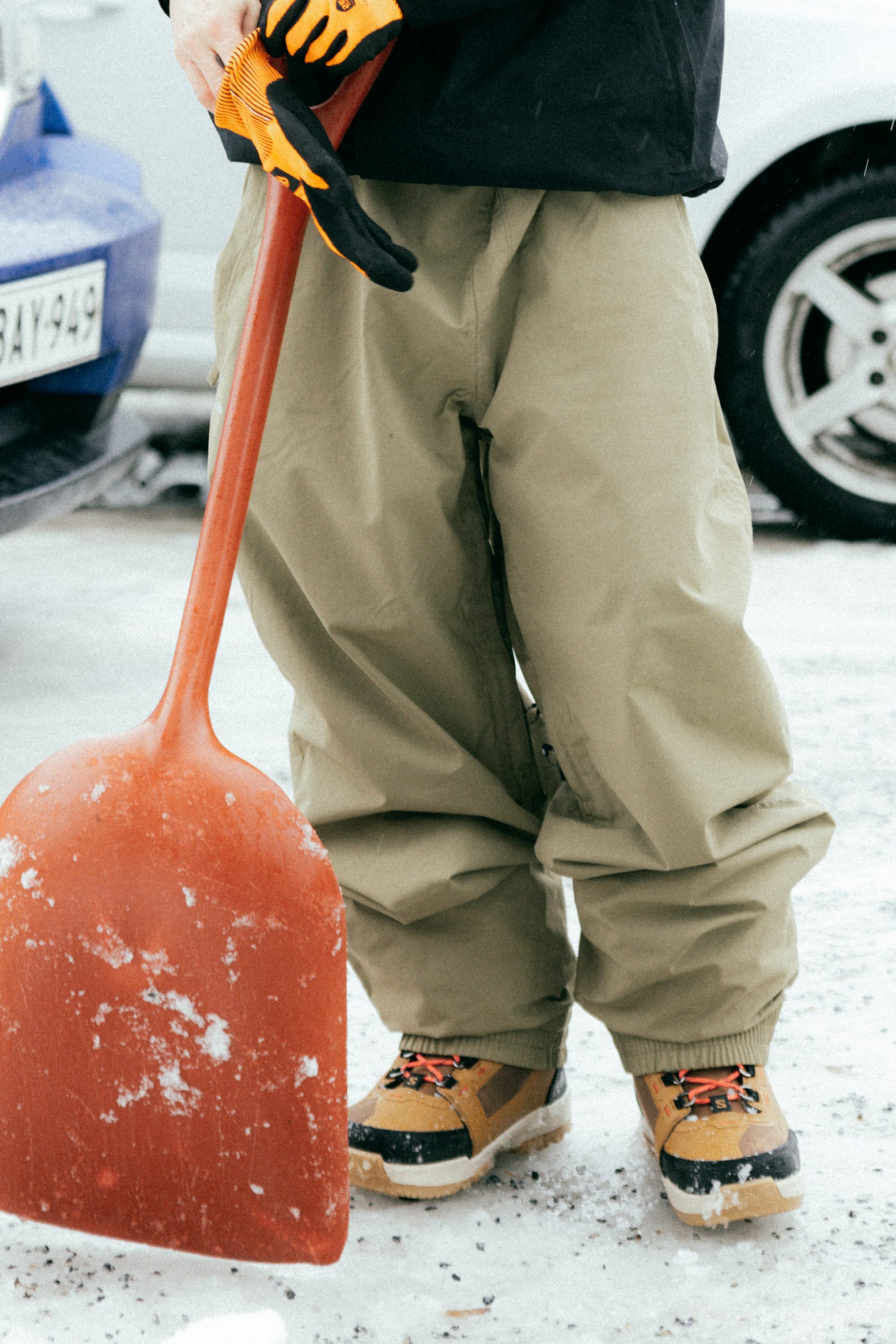A person in tan boots, orange gloves, and 686 Dojo® GORE-TEX Pants stands on snow holding a large red snow shovel.