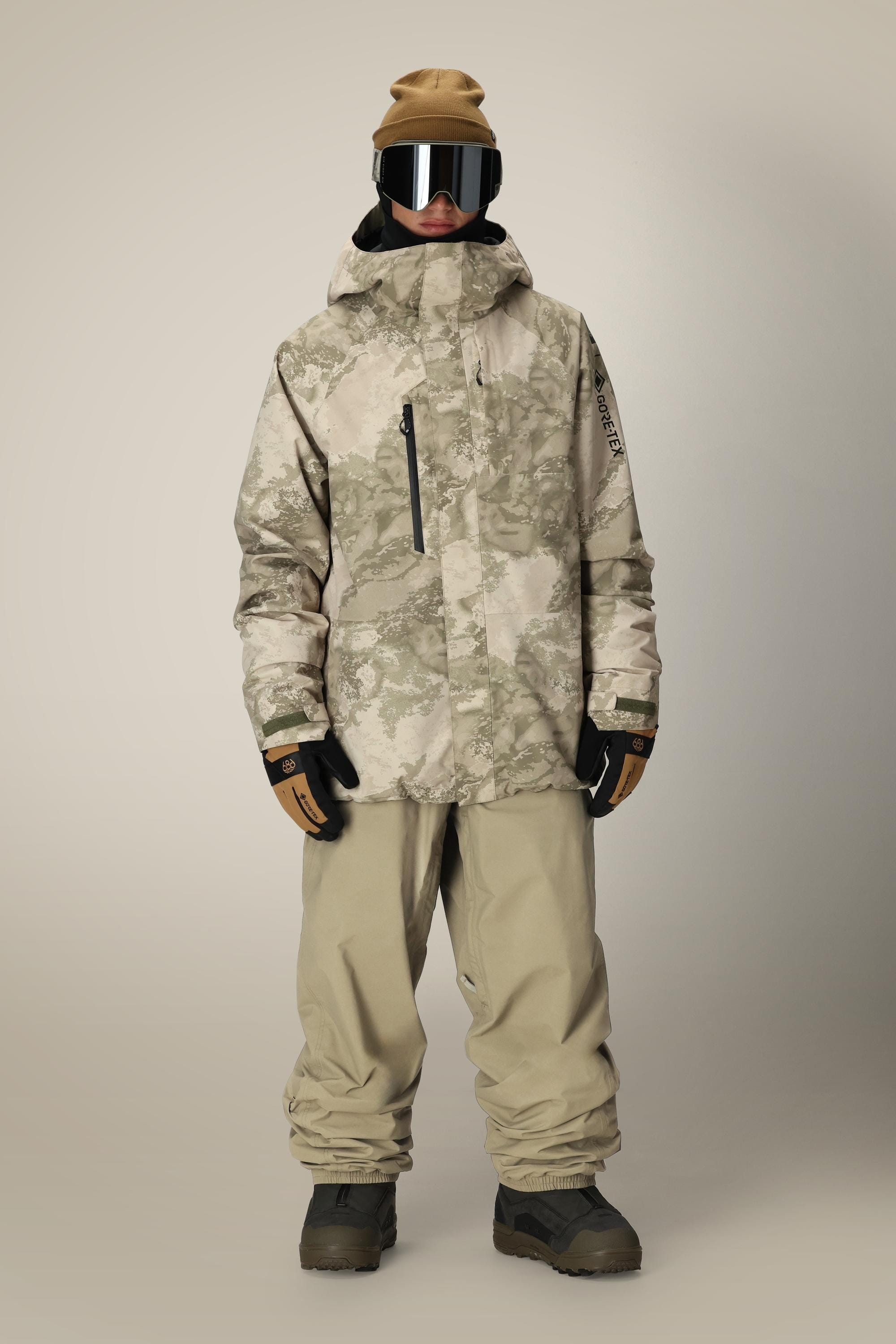 A person stands facing forward wearing a tan beanie, black goggles, a camo-patterned winter jacket, 686 Dojo® GORE-TEX Pant by 686, brown gloves, and dark boots against a plain light background.