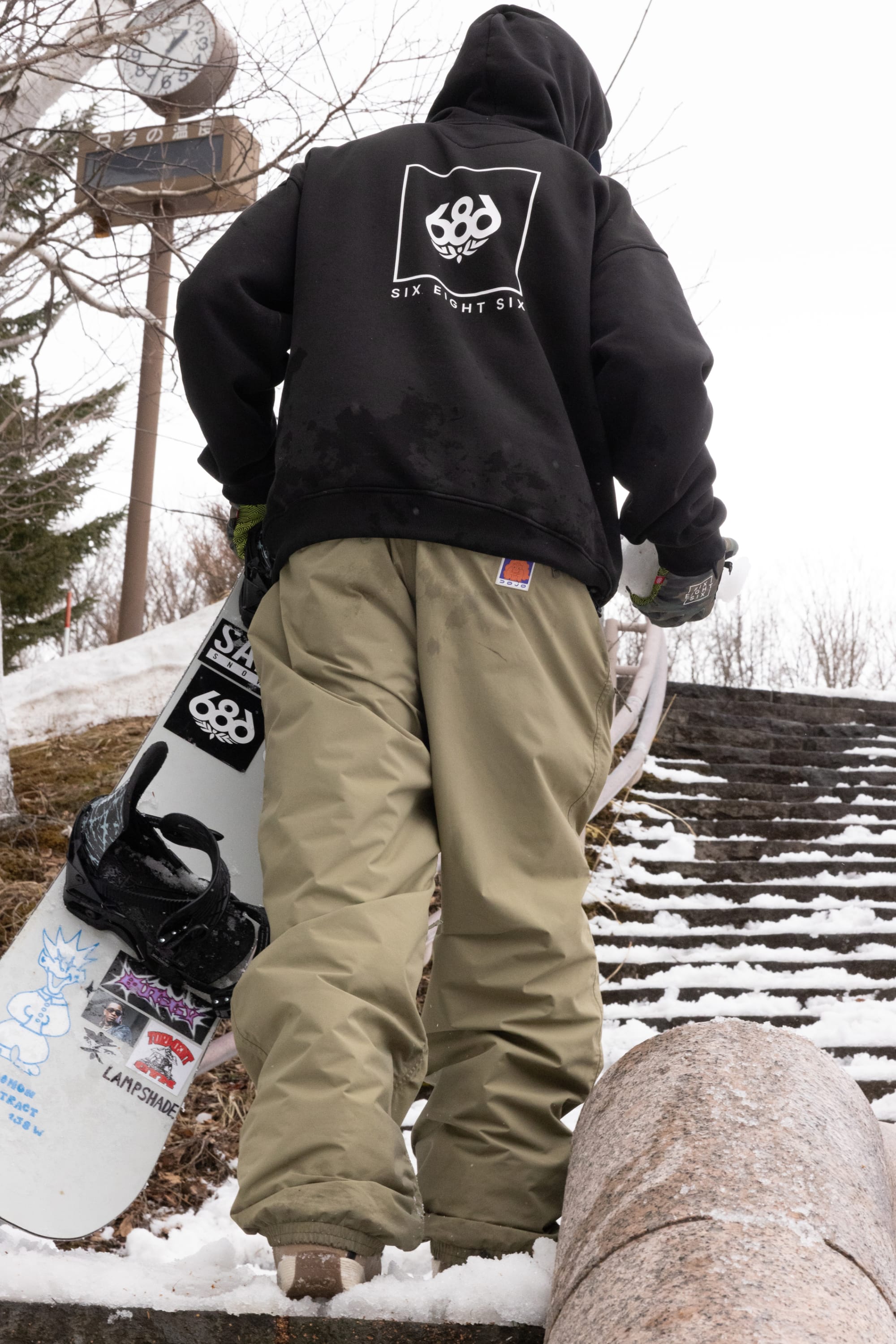 Wearing a black hoodie and 686 Dojo® GORE-TEX Pant, a person carries a snowboard up snow-dusted outdoor steps, with a clock visible on a post in the background.