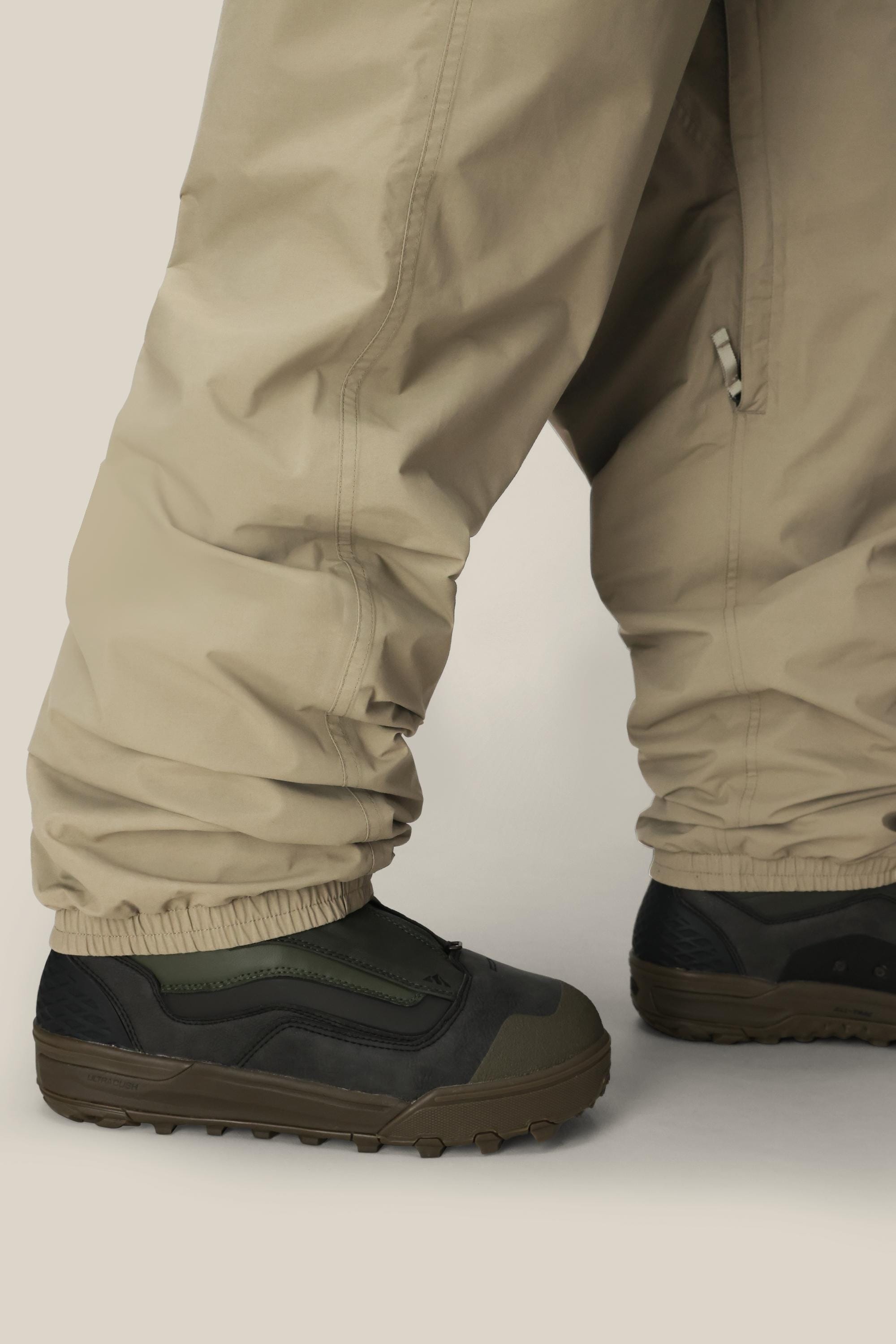 Close-up of a person wearing 686 Dojo® GORE-TEX Pants by 686, featuring a loose beige design draped over dark green and black rugged shoes, set against a neutral background.
