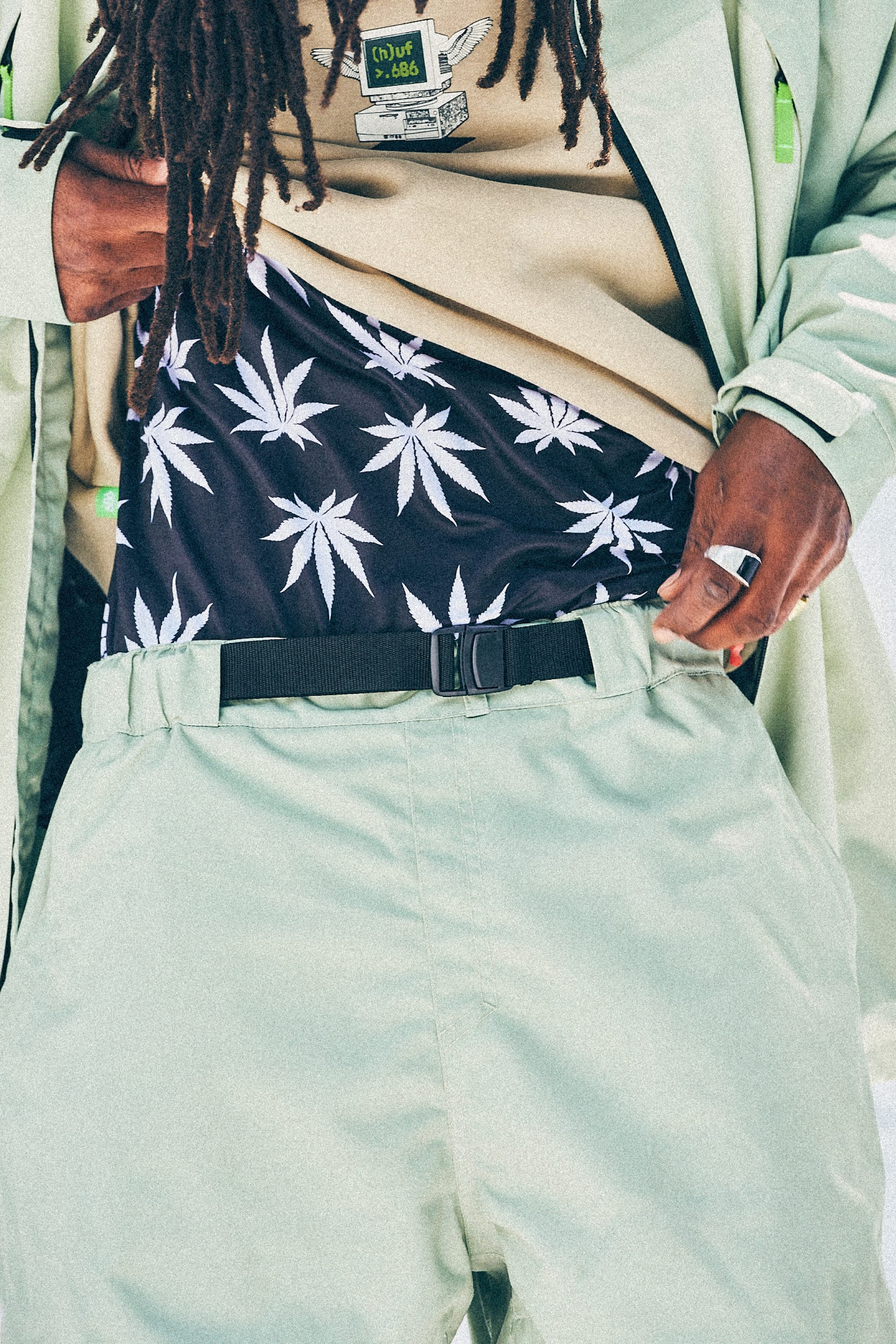 Wearing the 686 Mens HUF Double H Shell Pant in light green, paired with a matching jacket, this person reveals a black tee with a white cannabis leaf pattern. Accessories include a ring and long dreadlocks for a distinctive look.