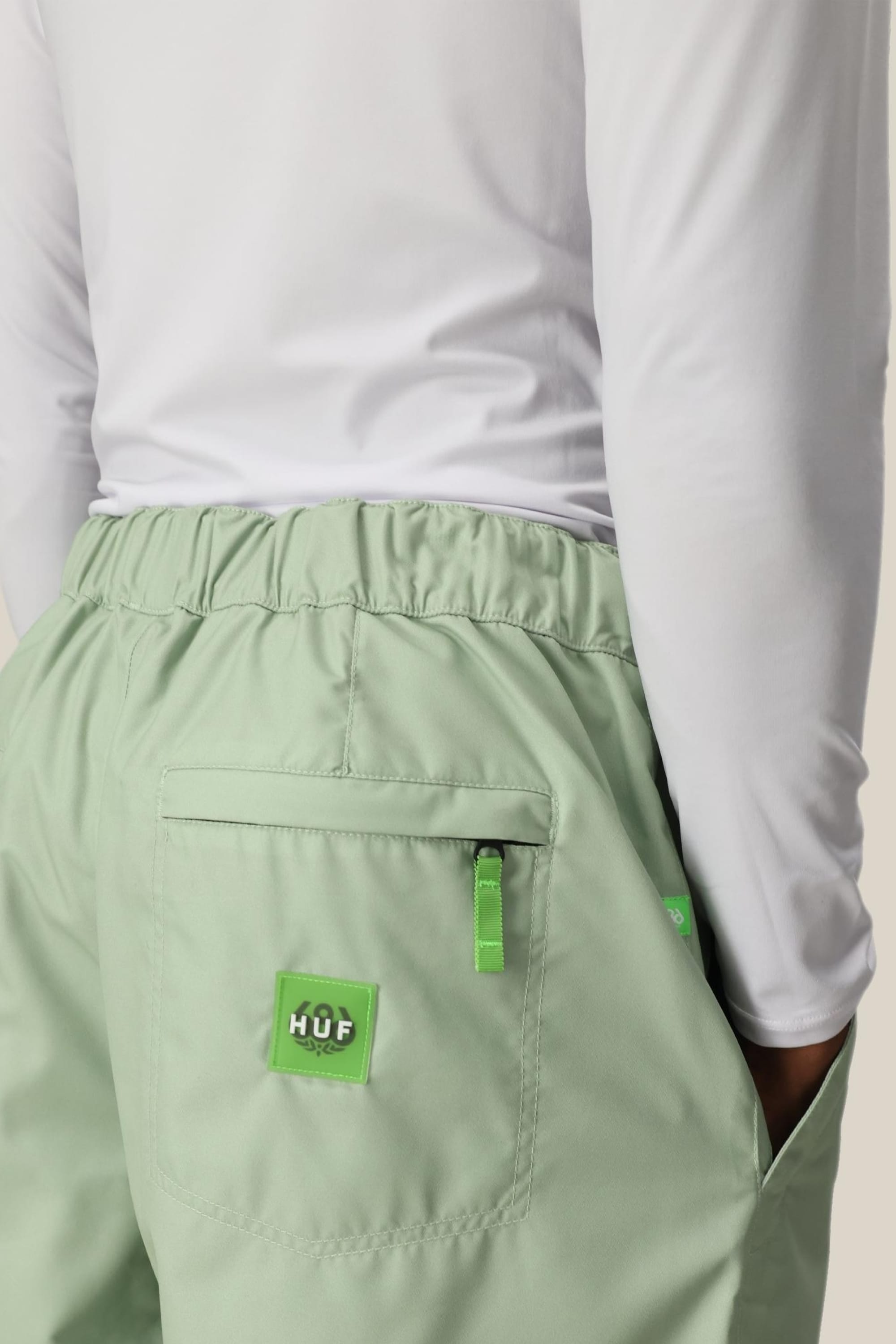 A person wearing 686 Mens HUF Double H Shell Pant by 686 in light green with a back pocket and green logo patch, styled with a white long-sleeve shirt. The sporty, casual polyester look is shown cropped at the waist and lower back.