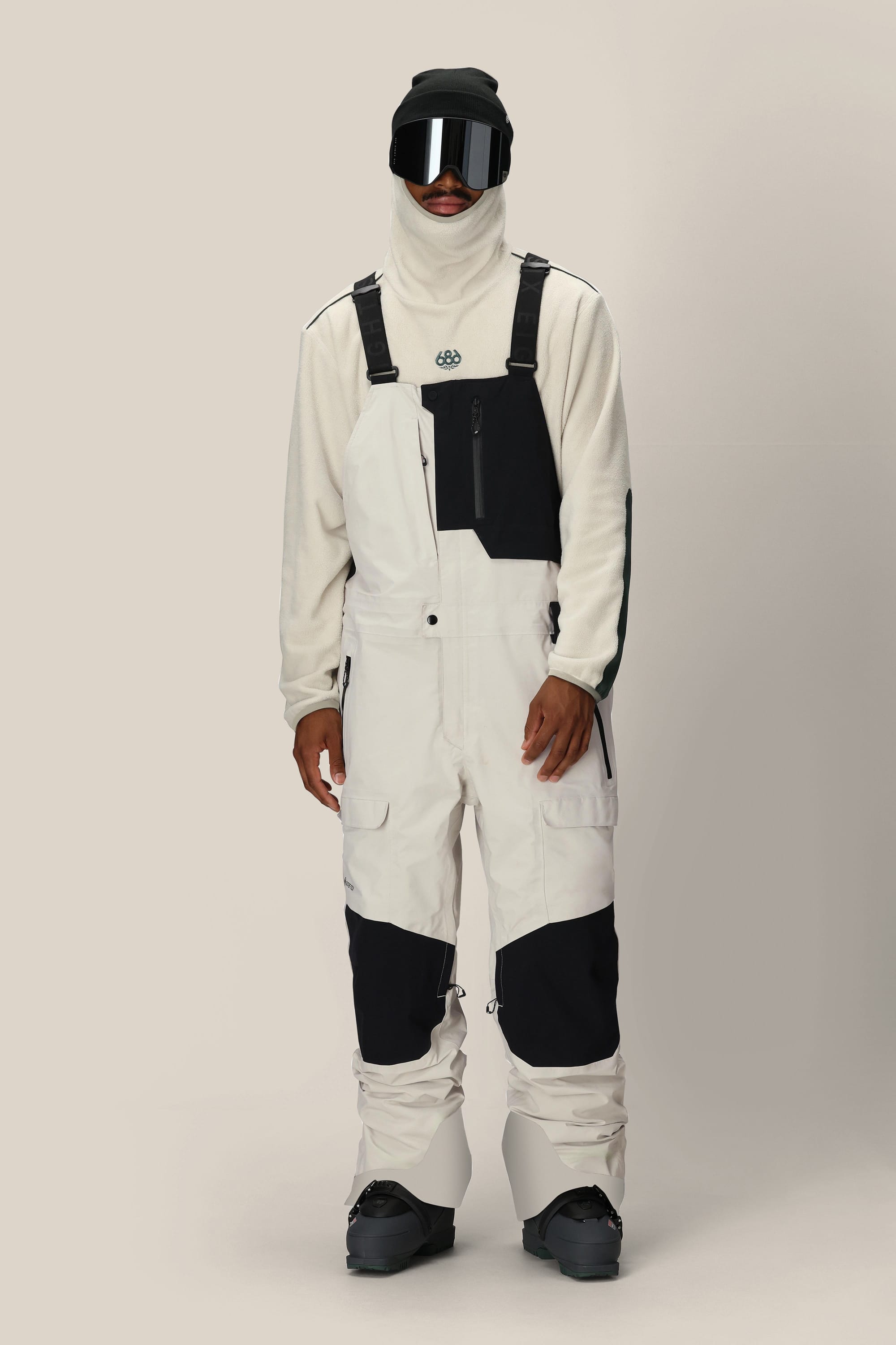Wearing the 686 Men's GORE-TEX Dispatch™ Shell Bib in black and white with a white fleece pullover, black snow boots, gloves, ski goggles, and a balaclava, a person stands against a plain light background.