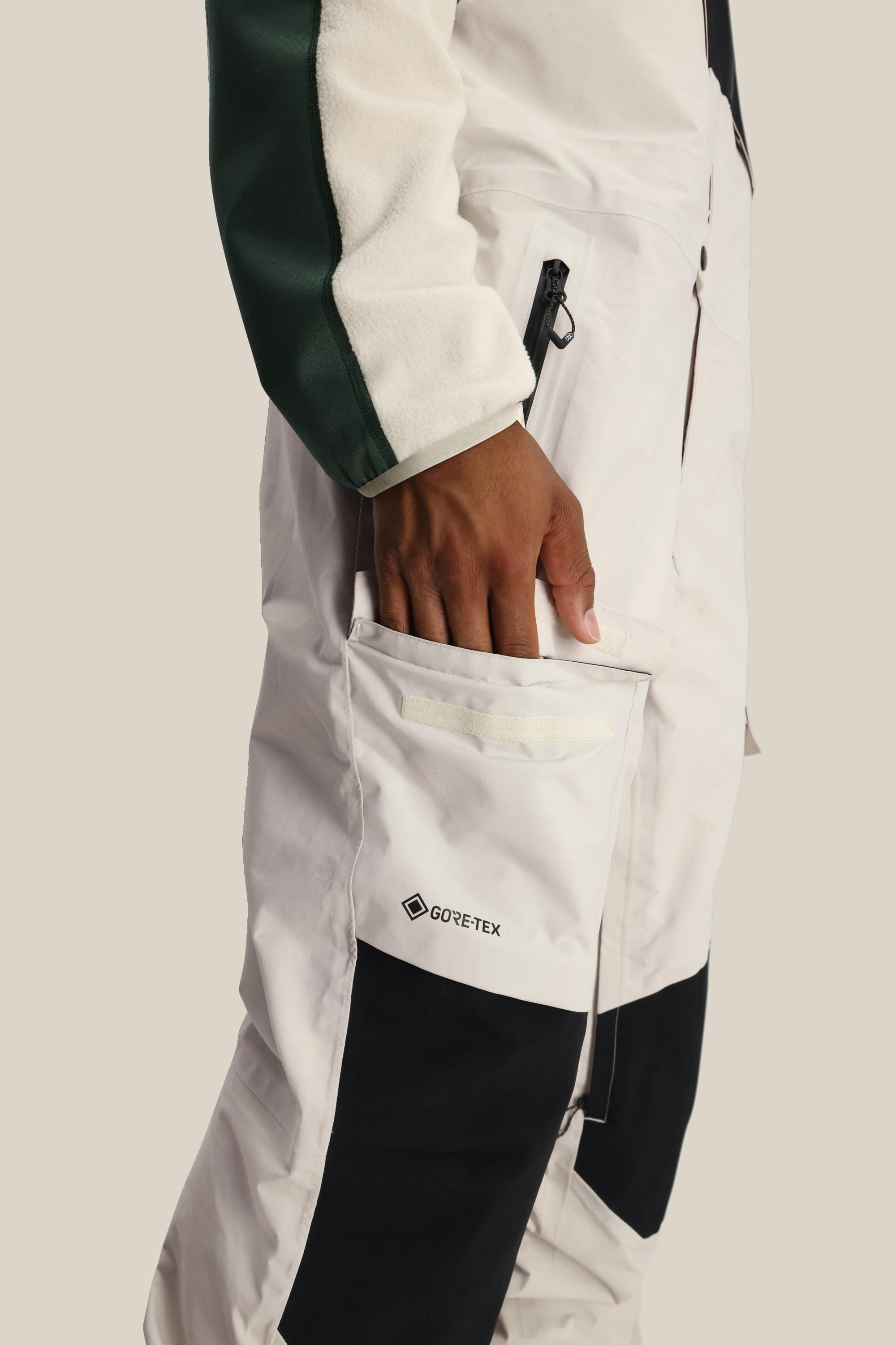 Wearing a white and black 686 GORE-TEX jacket with a green sleeve, a person places their hand into the zippered pocket of the 686 Men's GORE-TEX Dispatch™ Shell Bib against a plain light background.