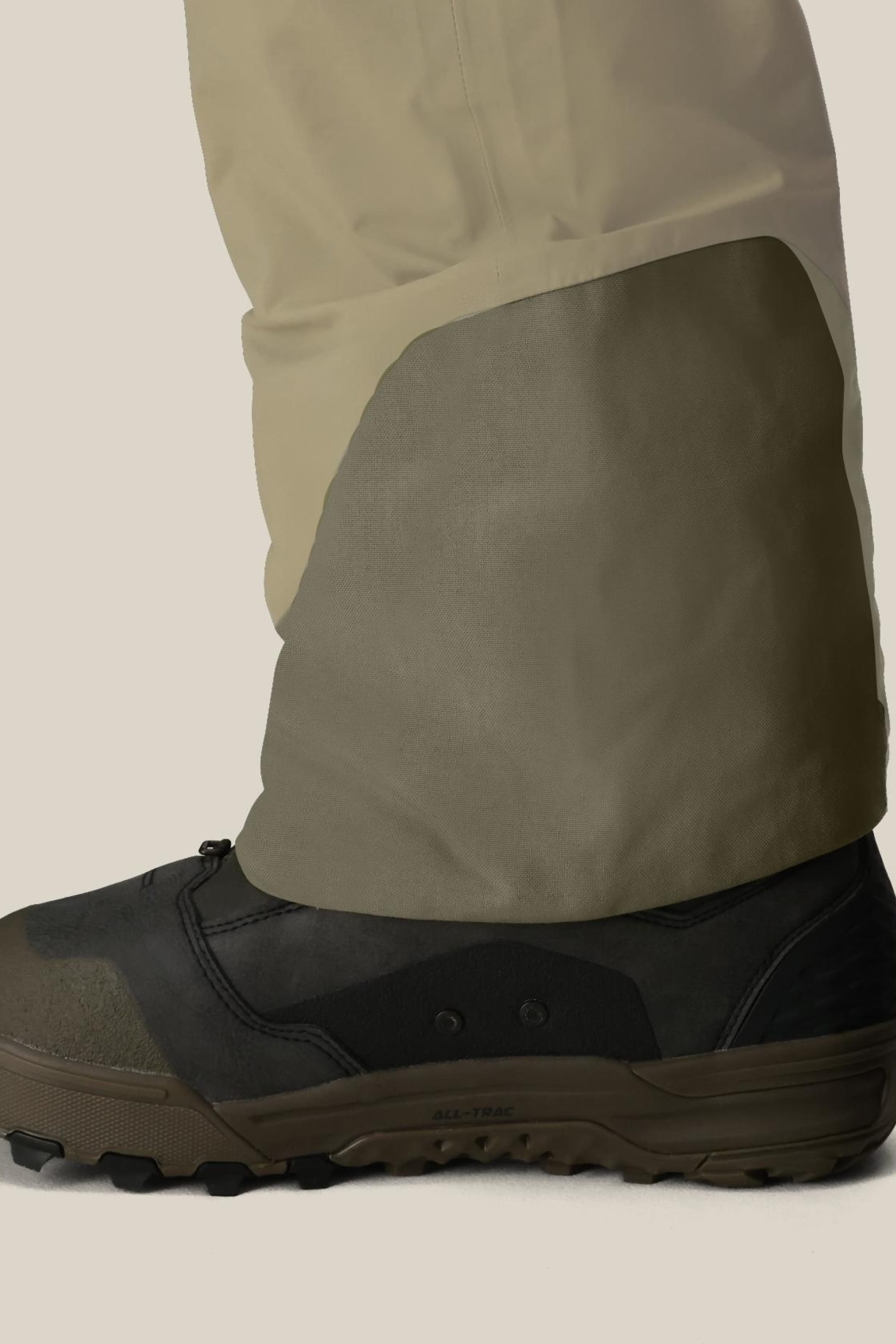 A close-up of a person's lower leg in tan pants with reinforced fabric over brown and black outdoor shoes, paired with the 686 Men's GORE-TEX Dispatch™ Shell Bib by 686, set against a light background.