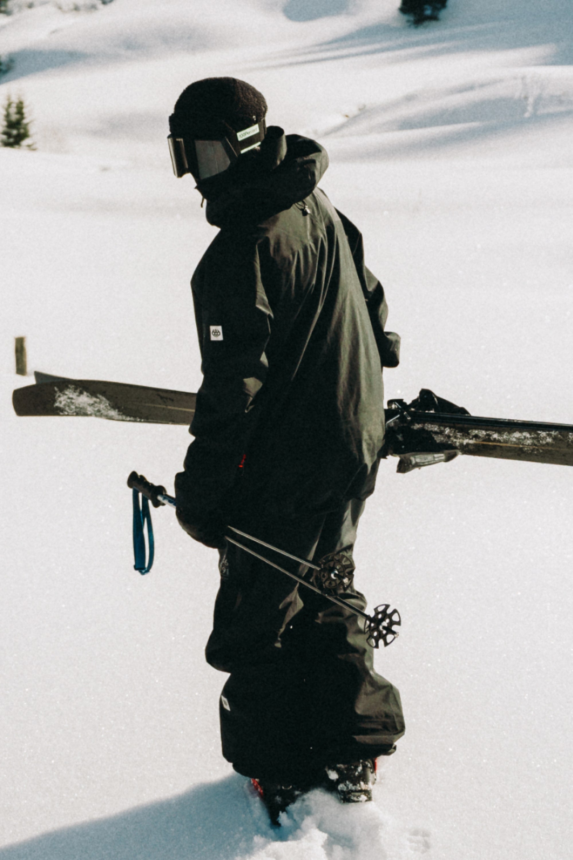 A person in black winter gear, wearing the 686 Mens GORE-TEX 3L ATV Bib and goggles, stands in deep snow holding skis and poles, facing away in a snowy landscape.