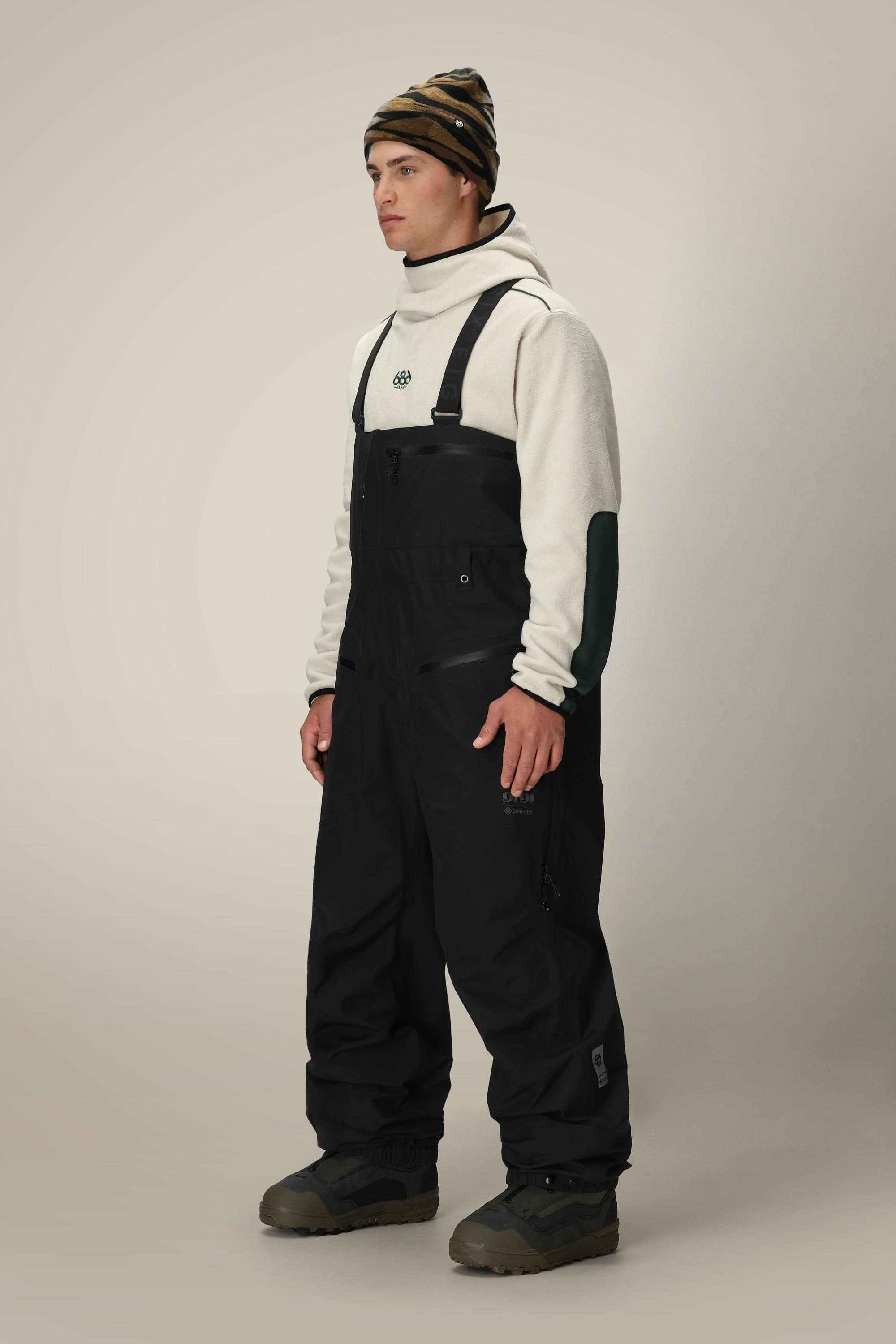 A person stands against a plain beige background wearing a white hoodie with black patches on the sleeves, 686 Mens GORE-TEX 3L ATV Bib snow overalls by 686, brown winter boots, and a striped beanie.