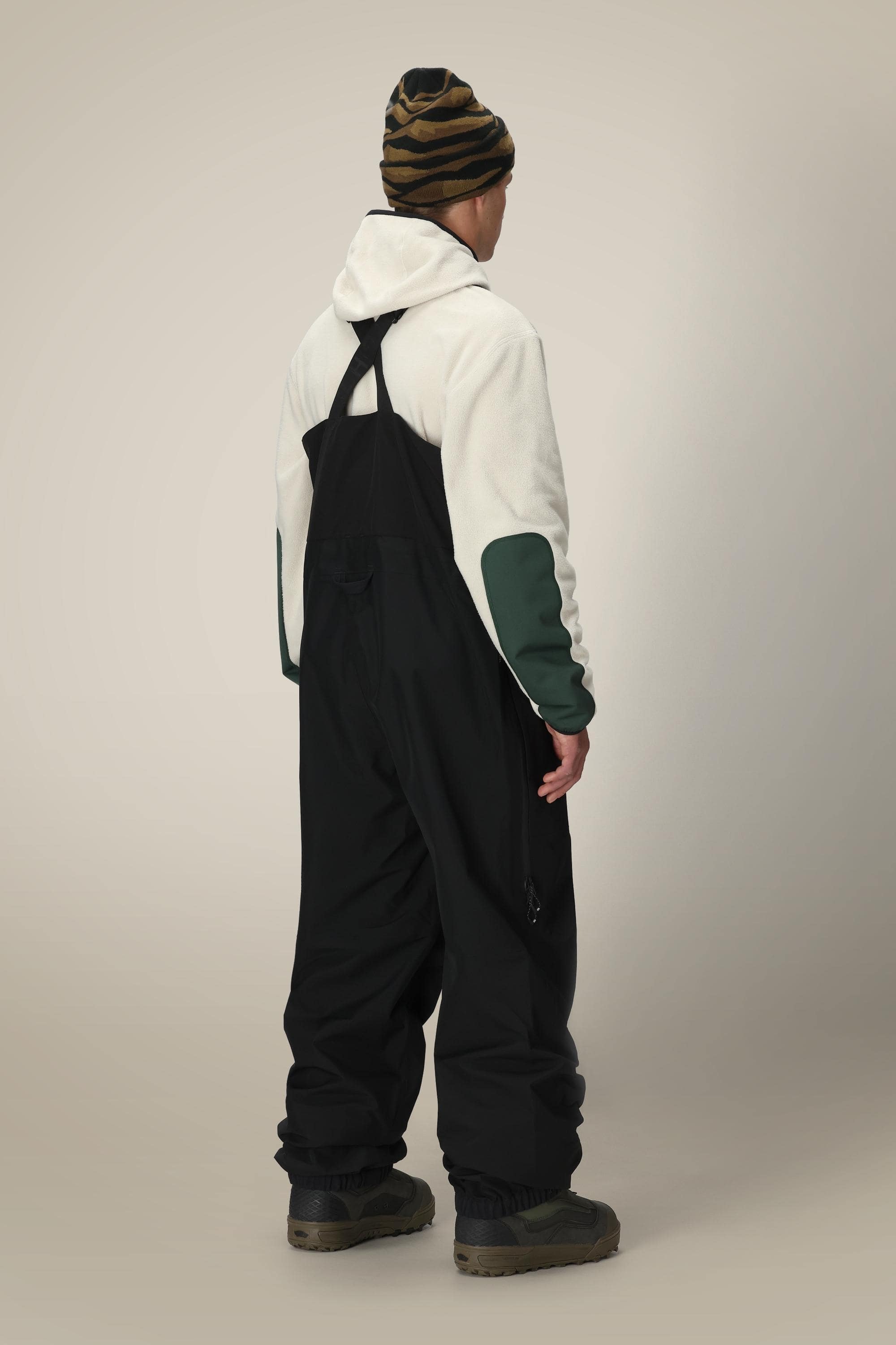 A person stands facing away, wearing the 686 Mens GORE-TEX 3L ATV Bib by 686, paired with a white and green hoodie, brown and black striped beanie, and dark boots against a plain light background.