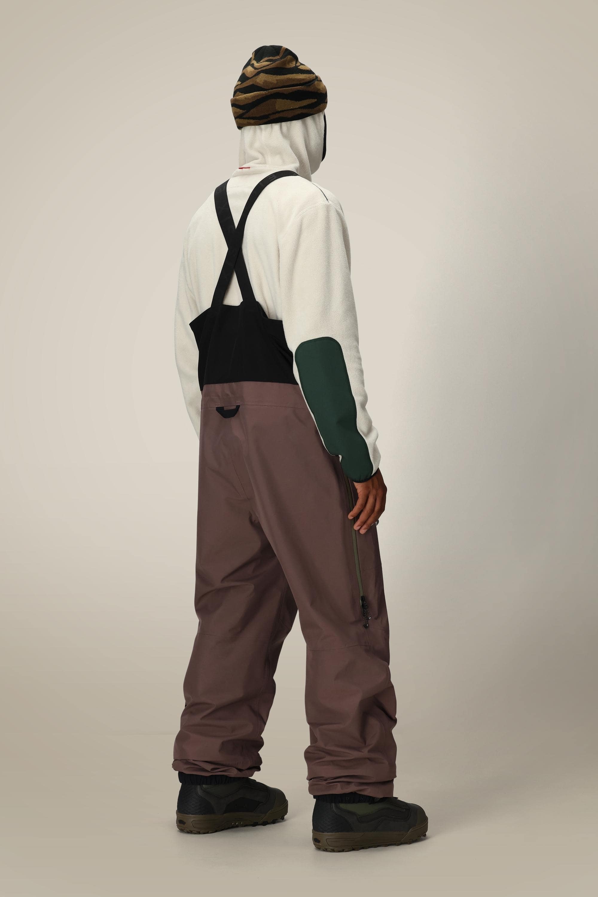 A person stands facing away, wearing a white hoodie with green sleeves, the 686 Mens GORE-TEX 3L ATV Bib by 686 with black suspenders, dark shoes, and a striped beanie hat against a plain light background.
