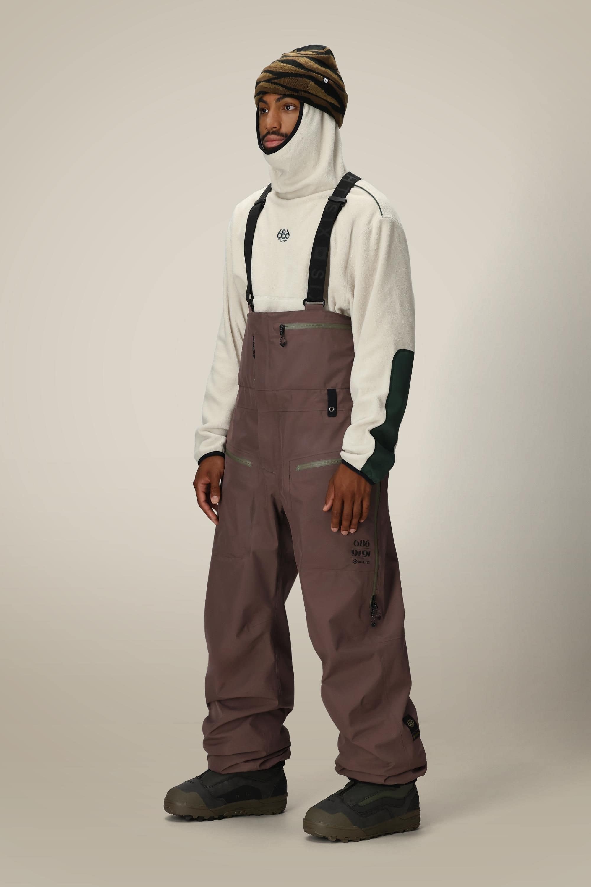A man stands against a plain light background wearing a beige balaclava, striped beanie, off-white fleece top with dark green accents, and loose-fitting 686 Mens GORE-TEX 3L ATV Bib snow pants with suspenders from 686.