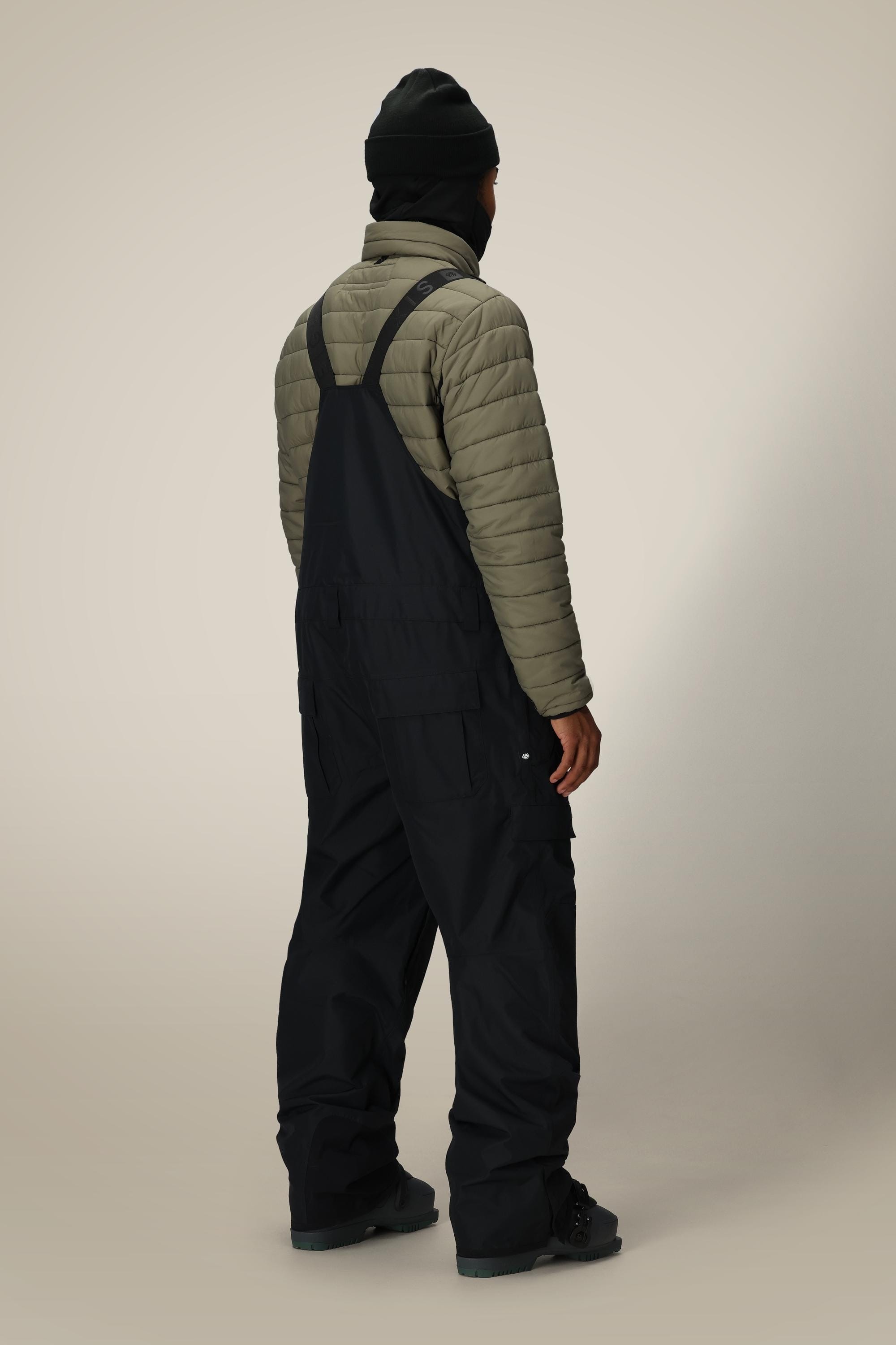 A person stands facing away against a plain beige background, wearing a black beanie, olive green quilted jacket, and the 686 Men's 20K Hot Lap™ Shell Bib by 686 in black with suspenders.