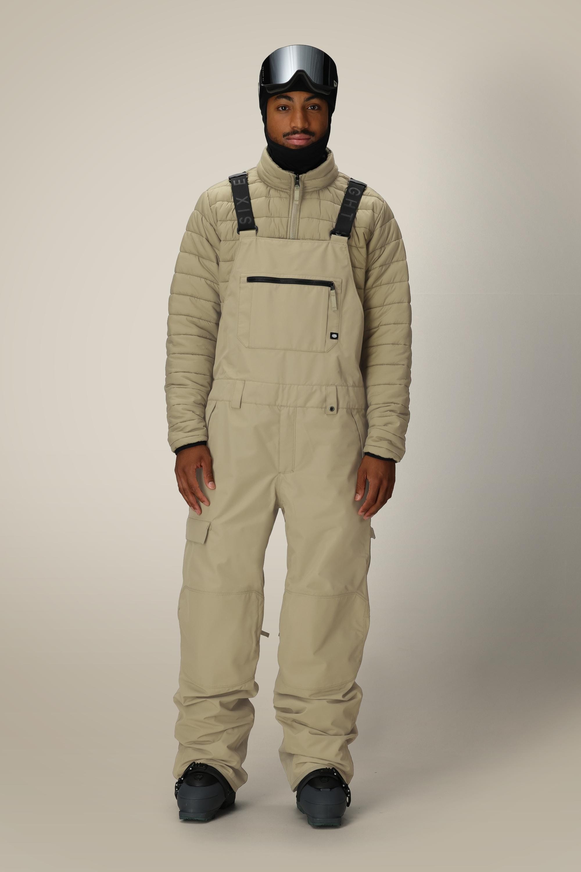 A person wears the 686 Men's 20K Hot Lap™ Shell Bib by 686 with a matching quilted jacket, black gloves, black ski boots, and a black ski helmet with goggles, standing against a plain light background.