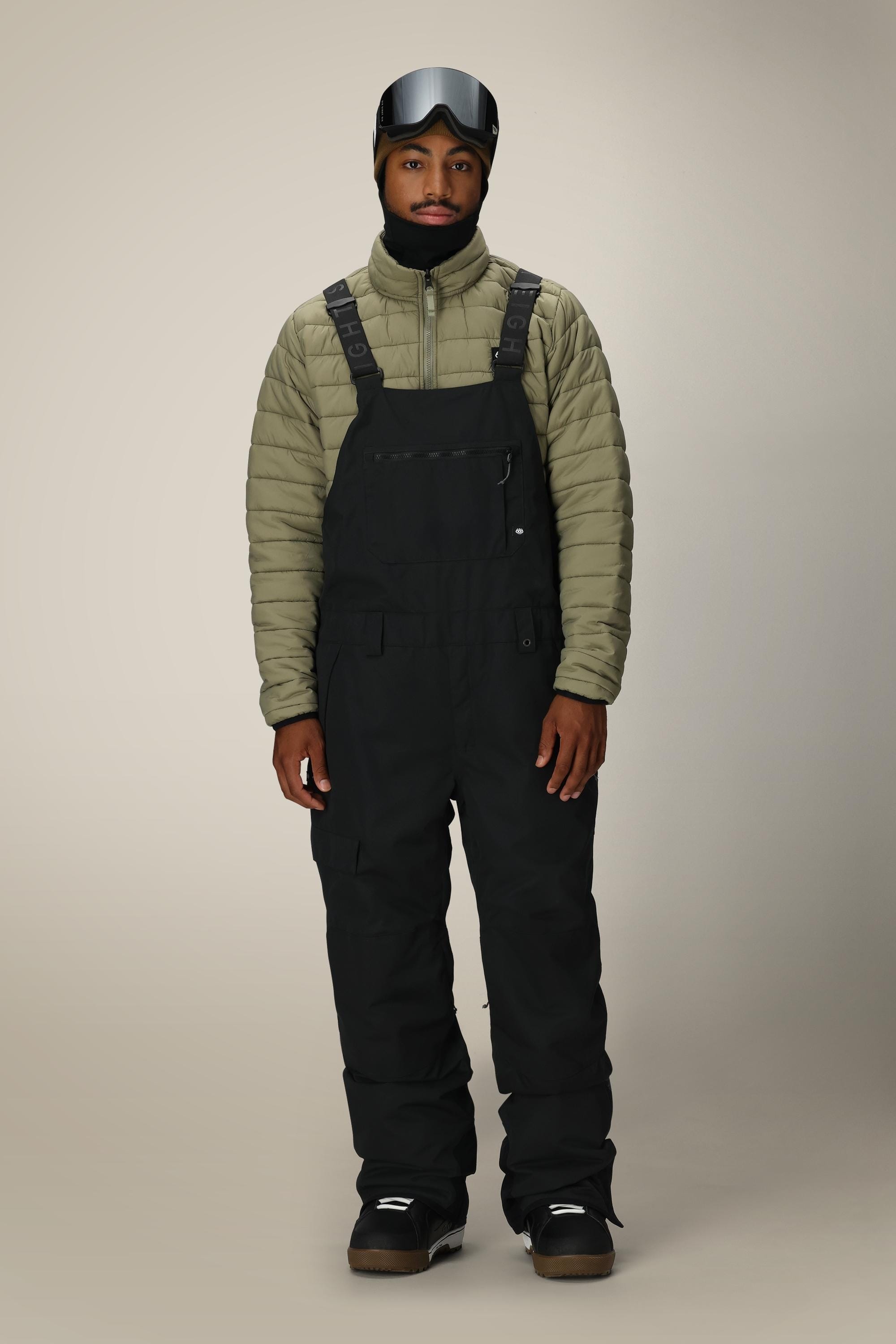 A person stands facing forward in the 686 Men's Hot Lap™ Insulated Bib by 686, paired with an olive green quilted jacket, black boots and gloves, and a ski helmet with goggles. The background is plain and light-colored.