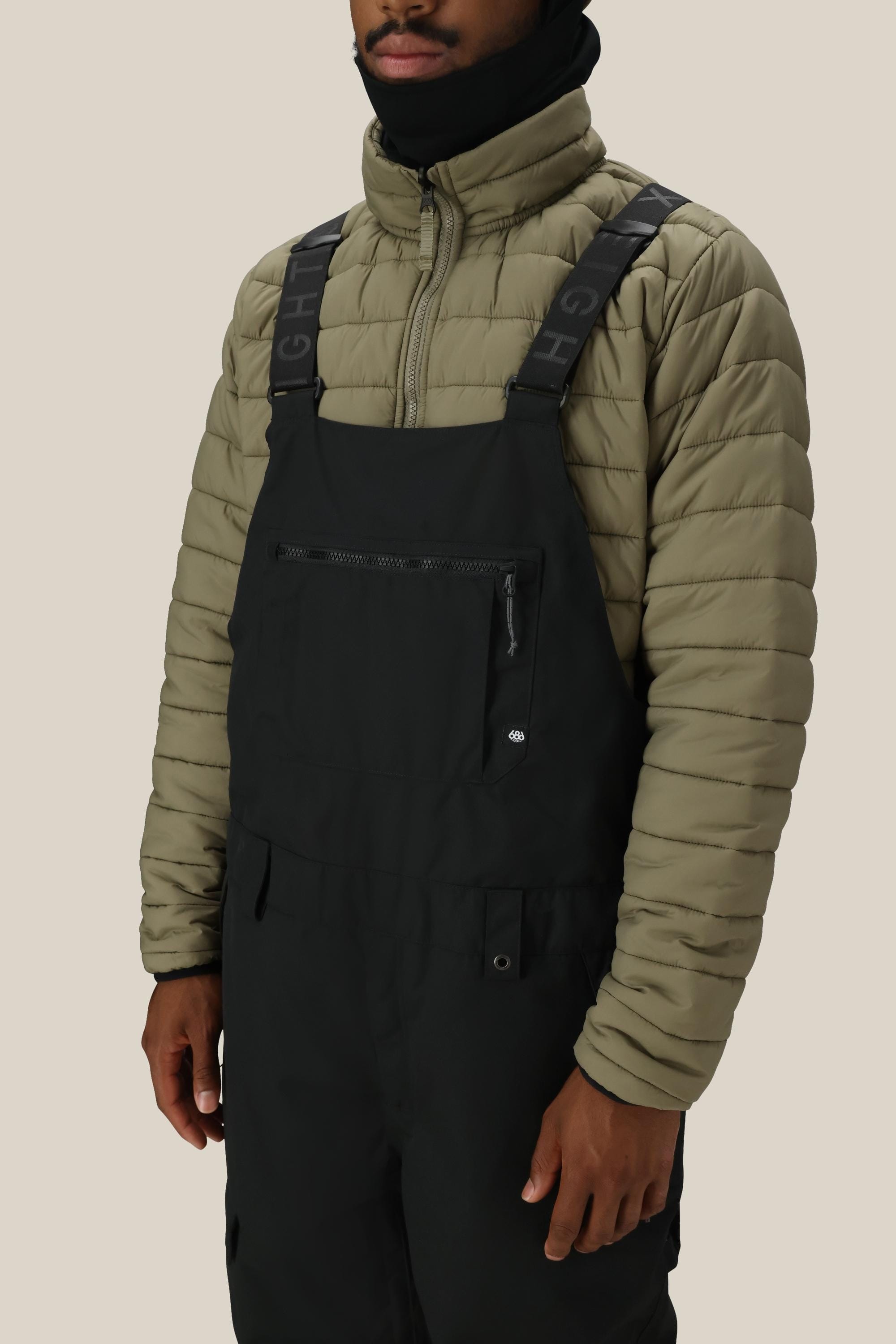 A person wearing the 686 Men’s Hot Lap™ Insulated Bib by 686, styled with a light olive puffer jacket, stands against a plain beige background.