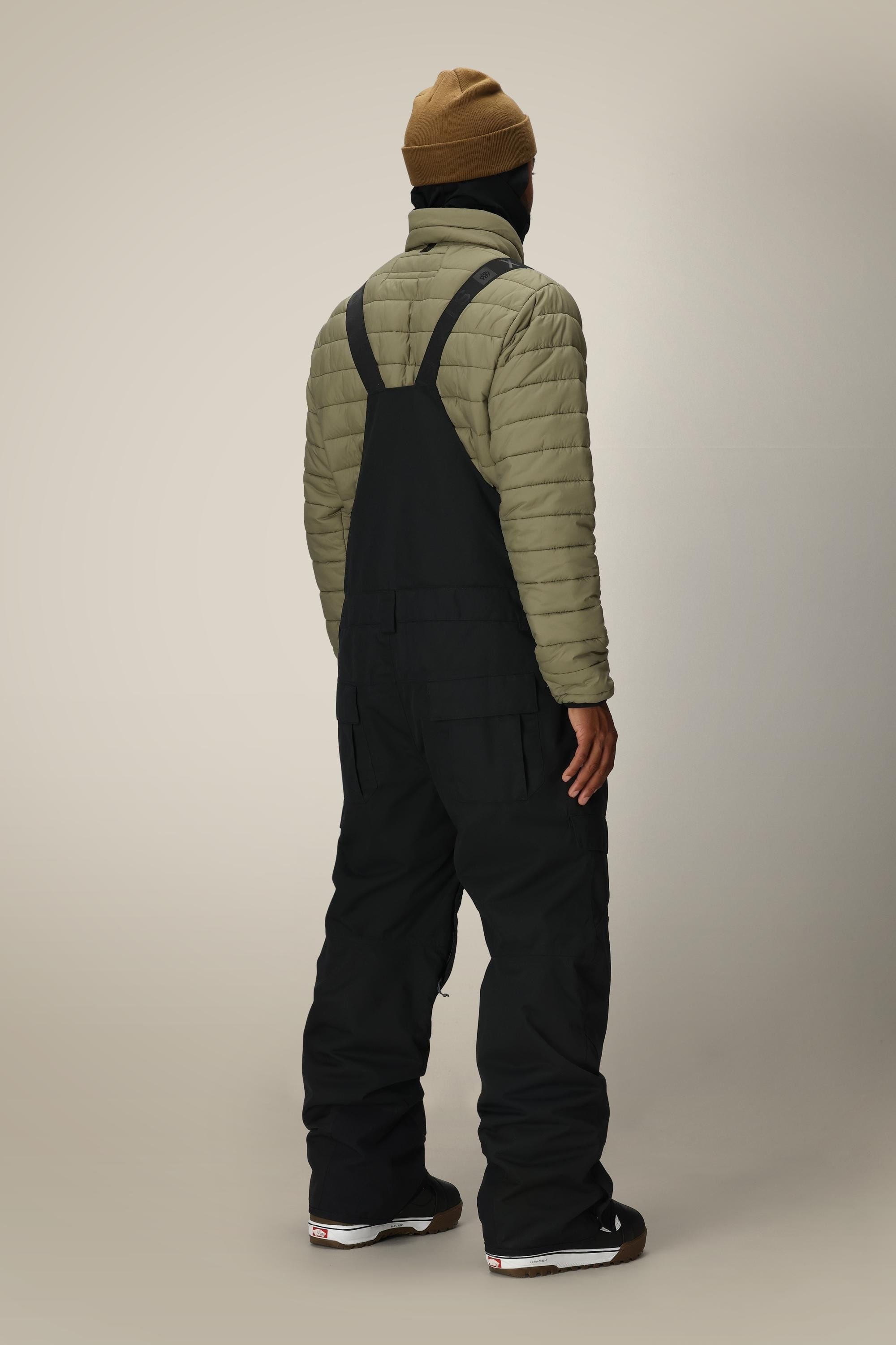 A person wearing a tan beanie, olive-green quilted jacket, and 686 Men's Hot Lap™ Insulated Bib stands facing away in a studio with a plain, light background.
