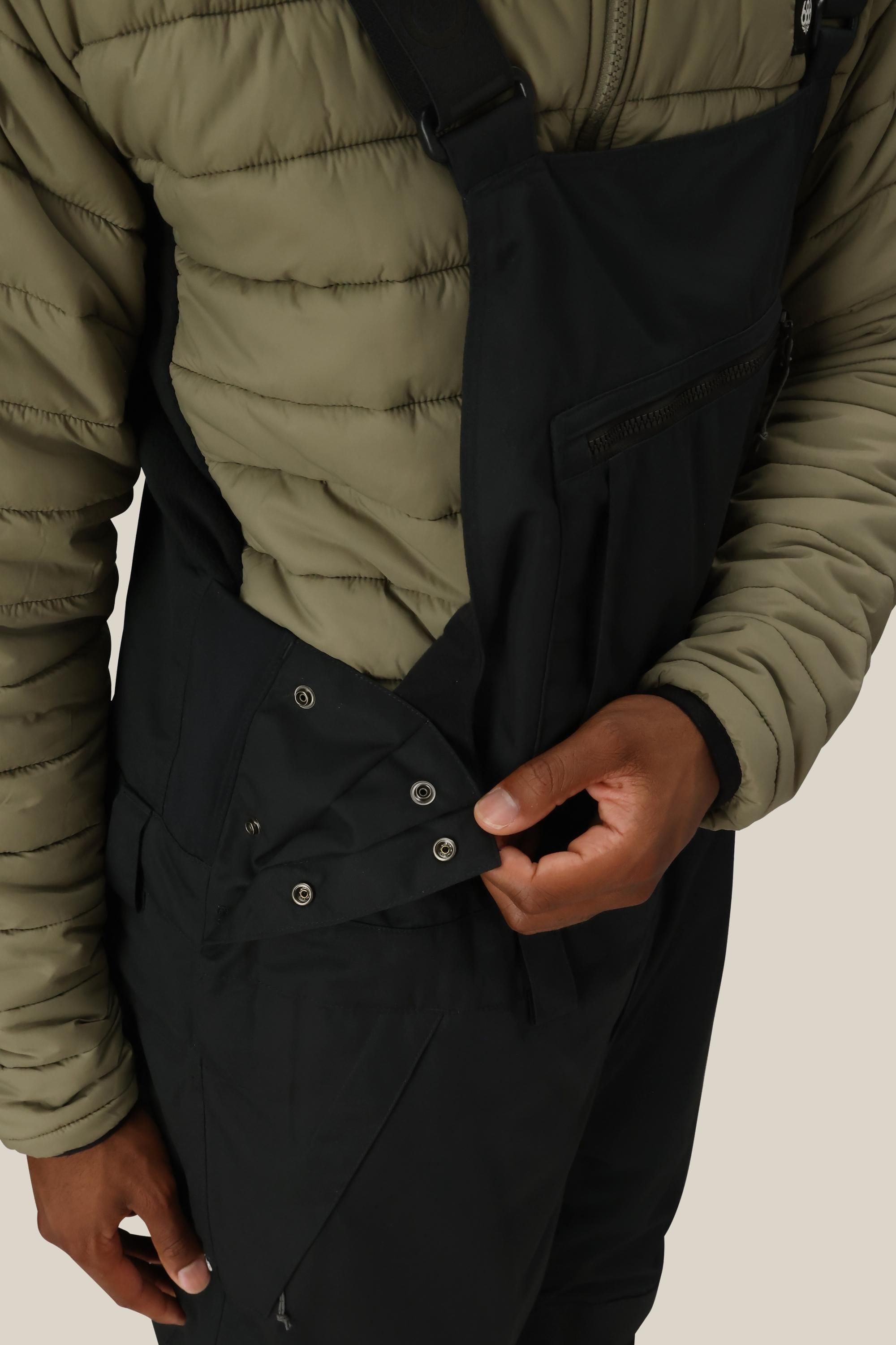 A person wearing a green quilted jacket and the 686 Men’s Hot Lap™ Insulated Bib in black opens a snap-button side flap on the bib, revealing the fastening detail.