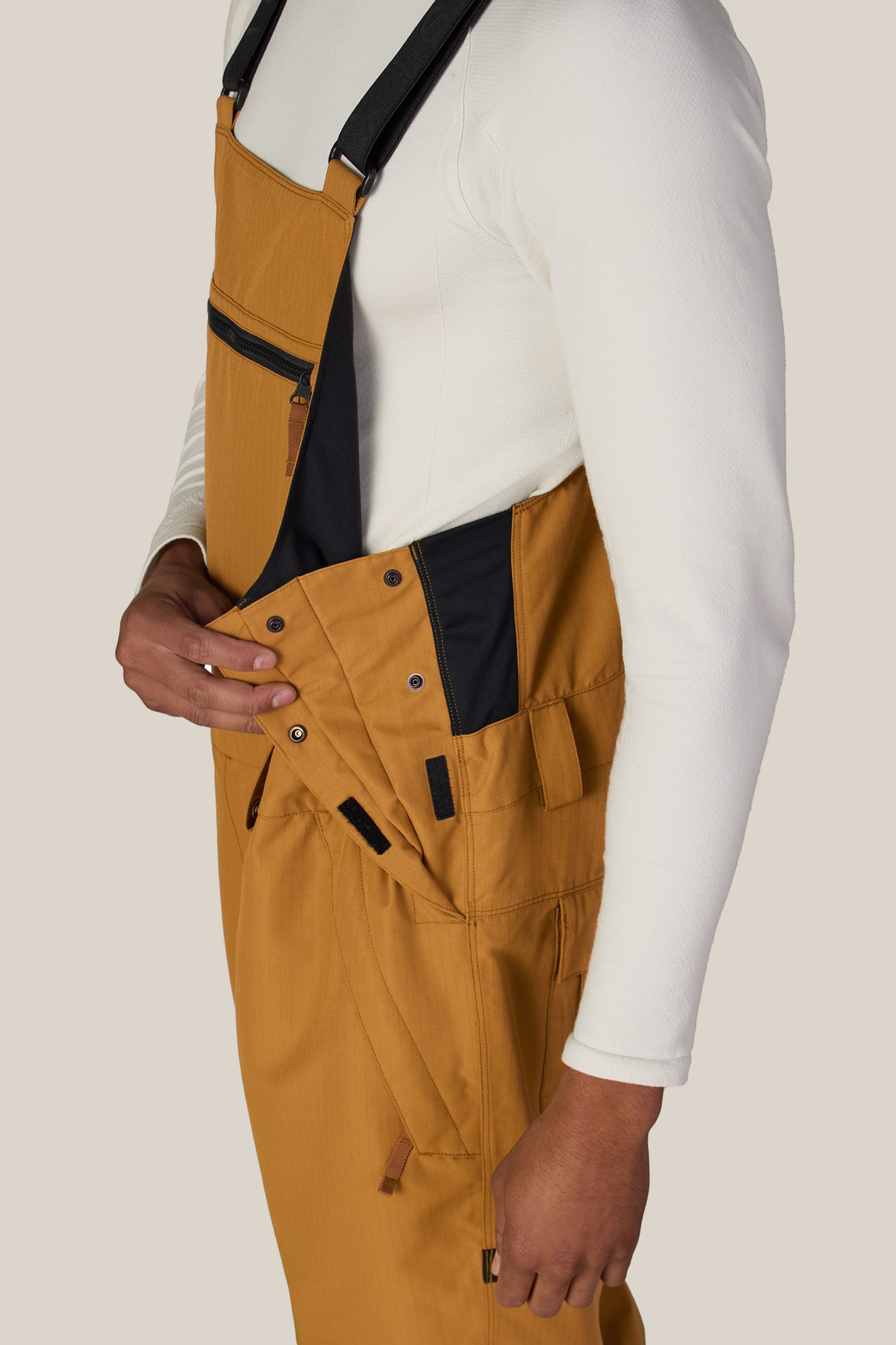 A person wearing the 686 Men’s Hot Lap™ Insulated Bib in mustard yellow over a white long-sleeve shirt adjusts the side buttons of the bib with one hand. The image is cropped at the shoulder and mid-thigh.