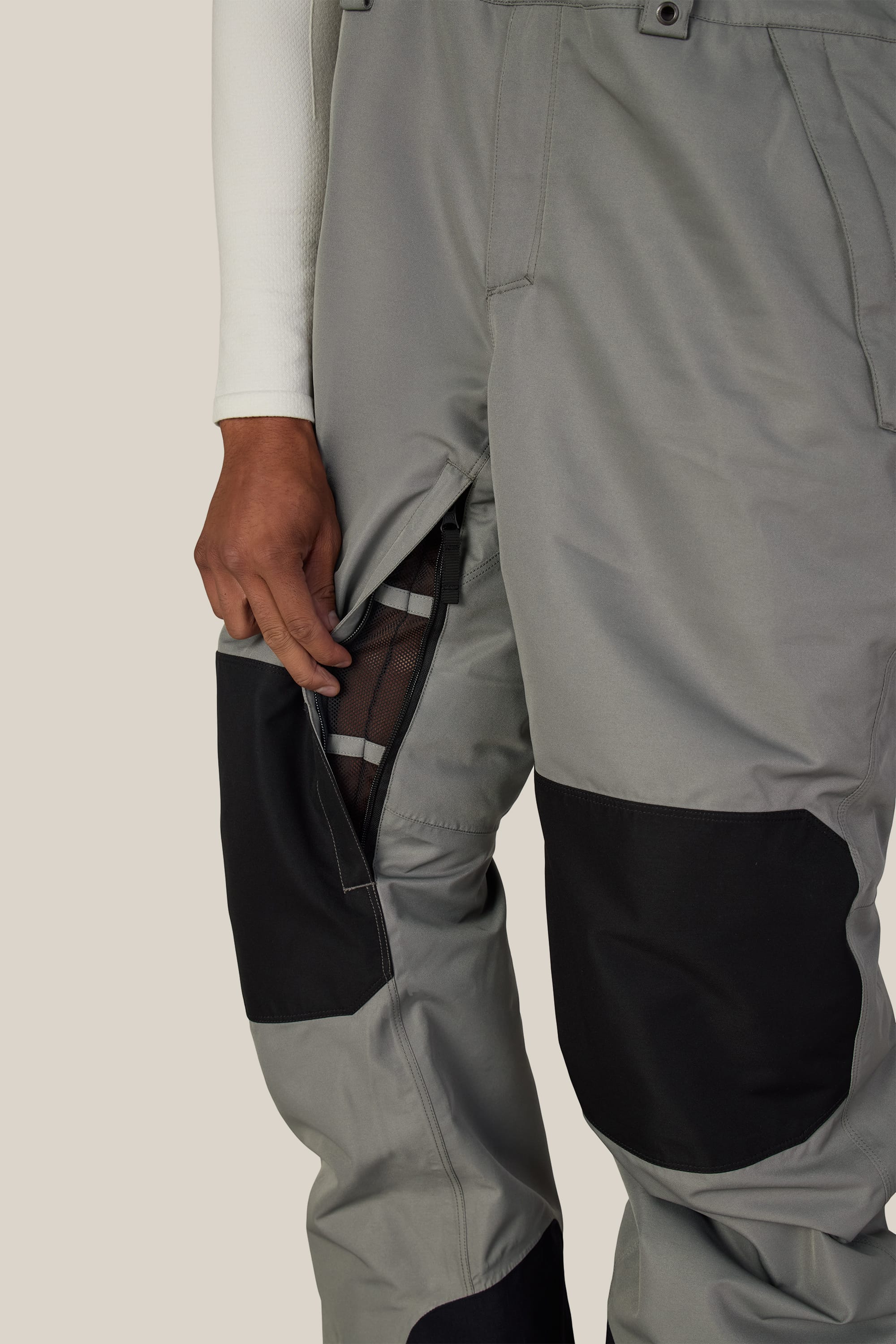 A person wearing the 686 Men’s Hot Lap™ Insulated Bib in gray and black unzips a thigh vent, revealing a mesh lining underneath. Only the lower half of their body is visible against a plain background.