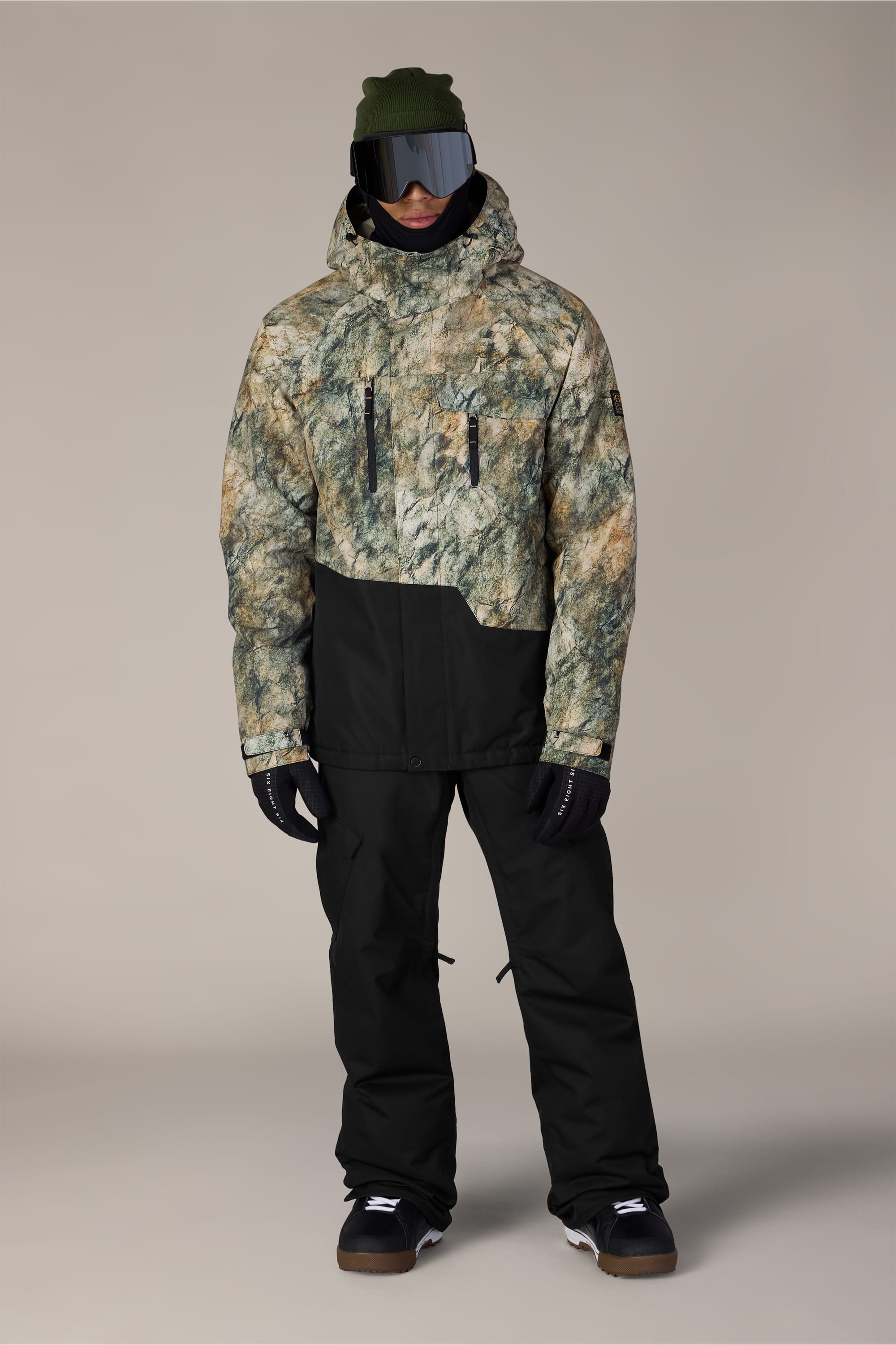 A person wearing a green and beige camo-patterned winter jacket, the 686 Men’s Infinity™ Insulated Cargo Pant by 686 in black, black gloves, boots, a green beanie, and large ski goggles stands against a plain background.