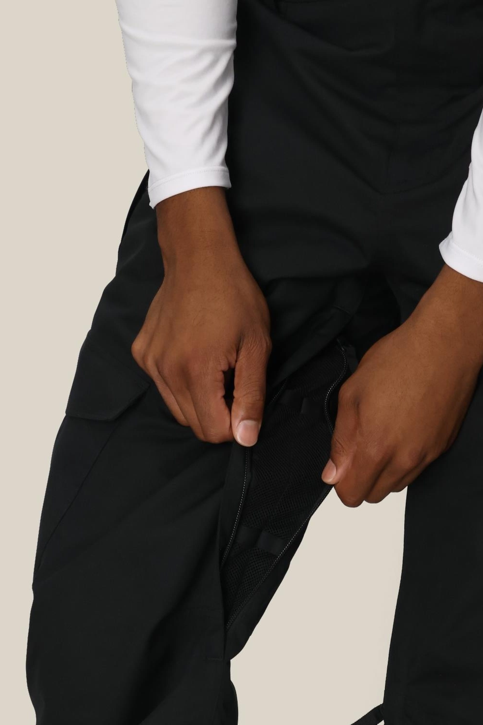 A person, torso and hands visible, unzips a thigh vent on 686 Men's Infinity™ Insulated Cargo Pant while wearing a white long-sleeve shirt.