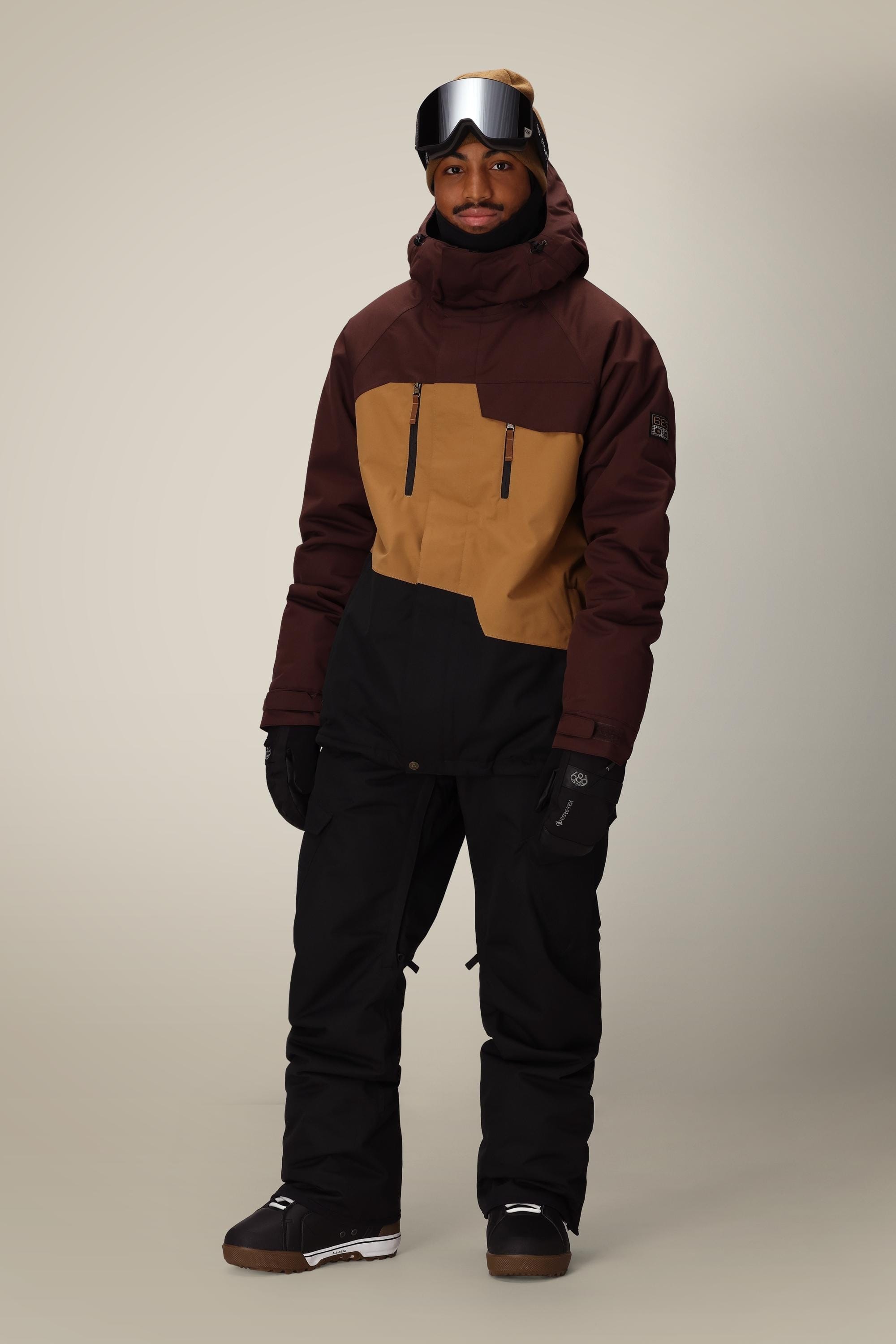 Facing forward against a light background, a person wears a brown and tan winter jacket, 686 Men's Infinity™ Insulated Cargo Pant by 686 in black, black gloves, brown boots, and reflective ski goggles.