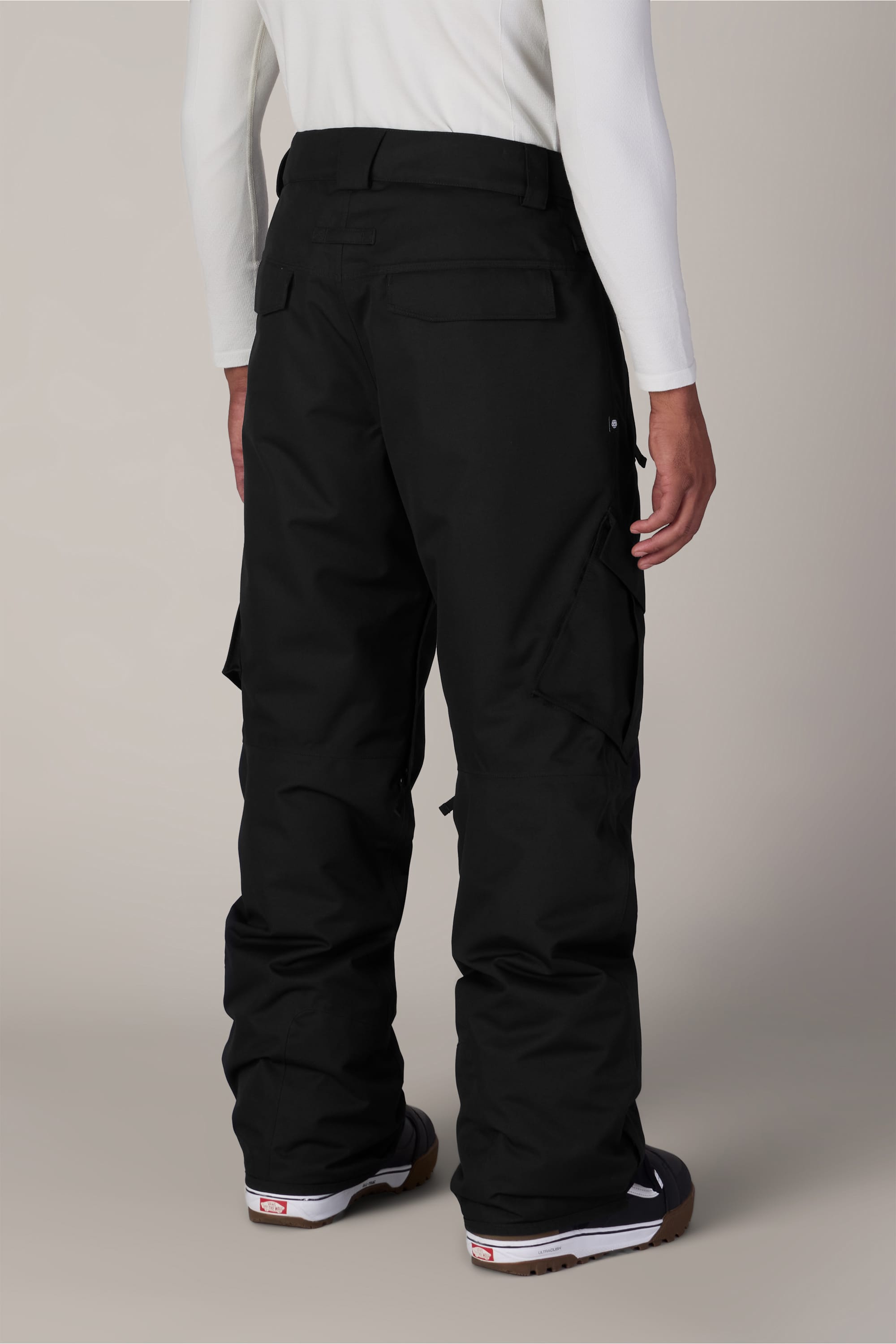 A person wearing the 686 Men’s Infinity™ Insulated Cargo Pant in black and a long-sleeve white shirt is photographed from behind. The pants by 686 feature large cargo pockets, and the individual is standing against a plain, light background.