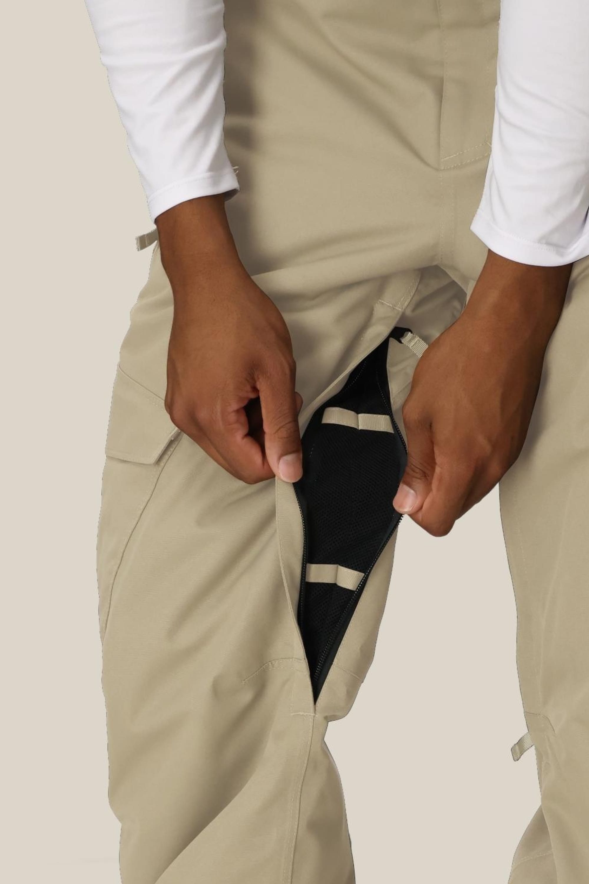 A person in a white long-sleeve shirt unzips the side vent of 686 Men's Infinity™ Insulated Cargo Pant in beige, revealing a black mesh lining. The background is plain and light-colored.